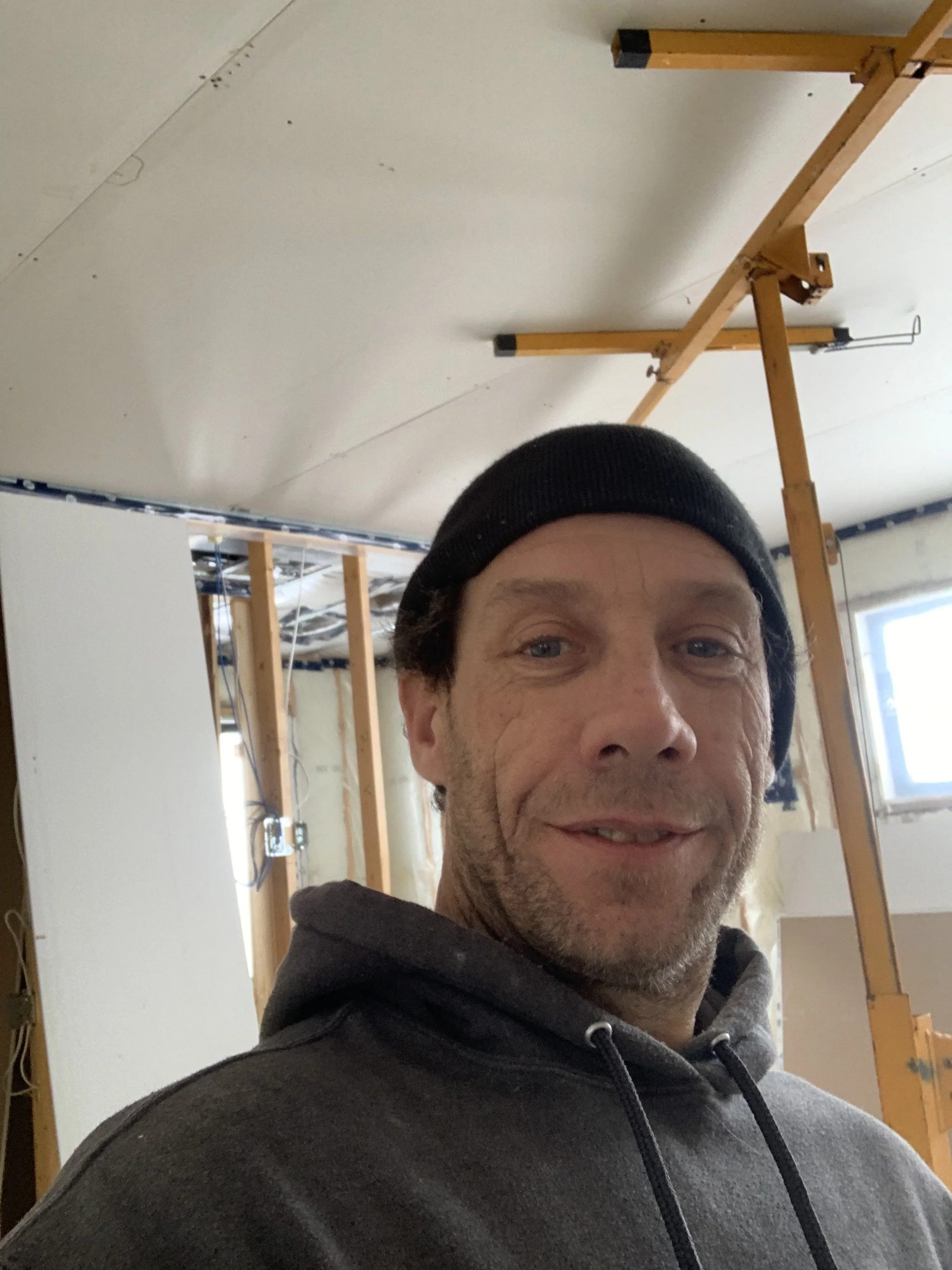 A man taking a selfie in a room under construction, with visible wooden framing and drywall installed on the ceiling.
