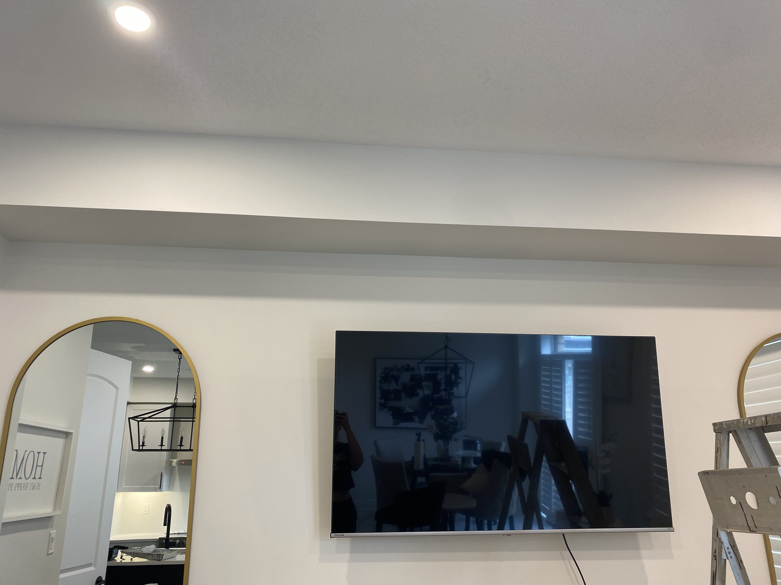 A living room with a wall-mounted television, a circular mirror, and a window with shutters. Part of a kitchen with a sink and faucet is visible in the background.