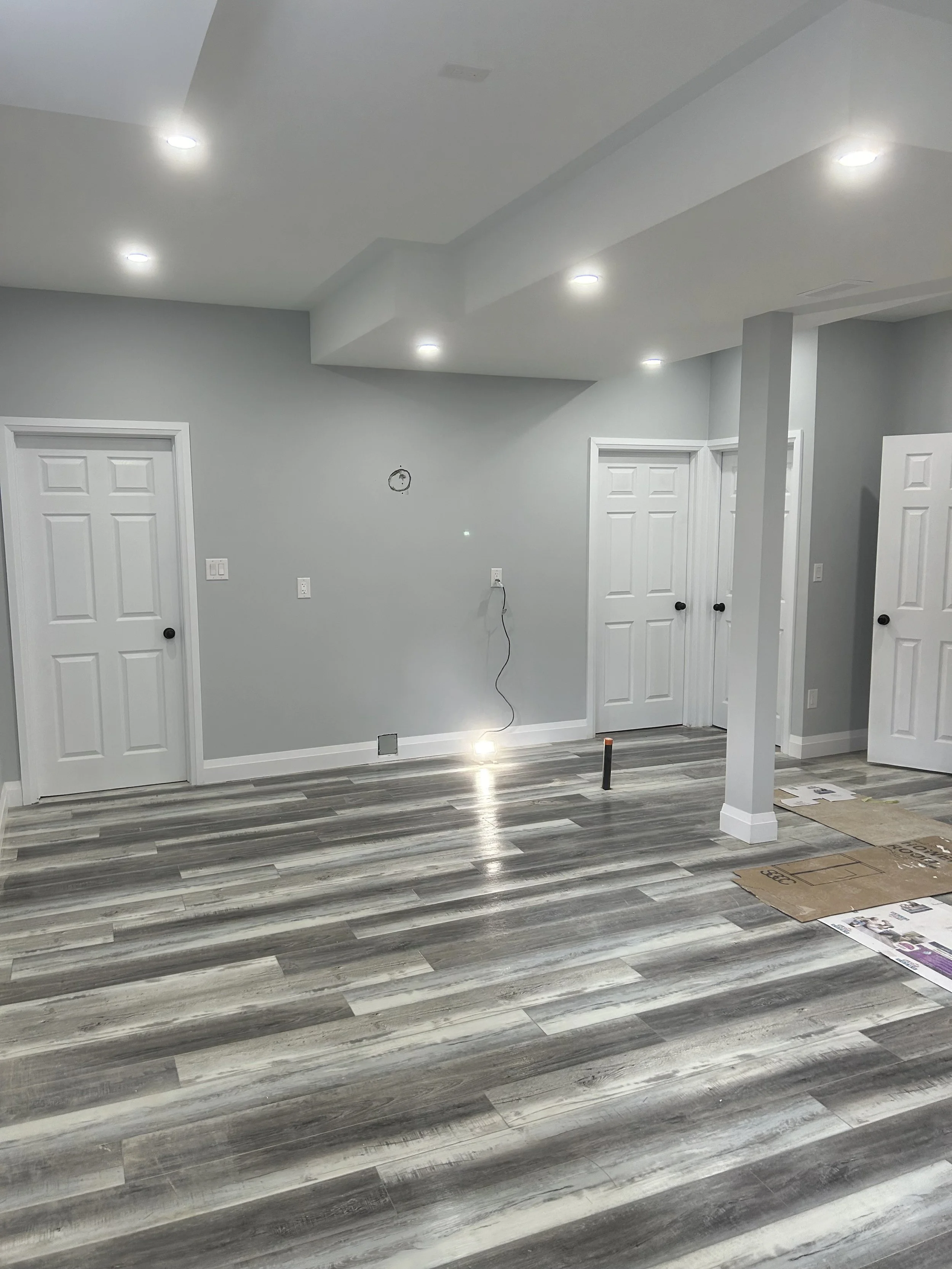 Interior of a room with light gray walls, white doors, and wood-patterned flooring. Recessed lighting fixtures are on the ceiling. A small hole with wires is on the wall, and there are some cardboard boxes and papers on the floor.