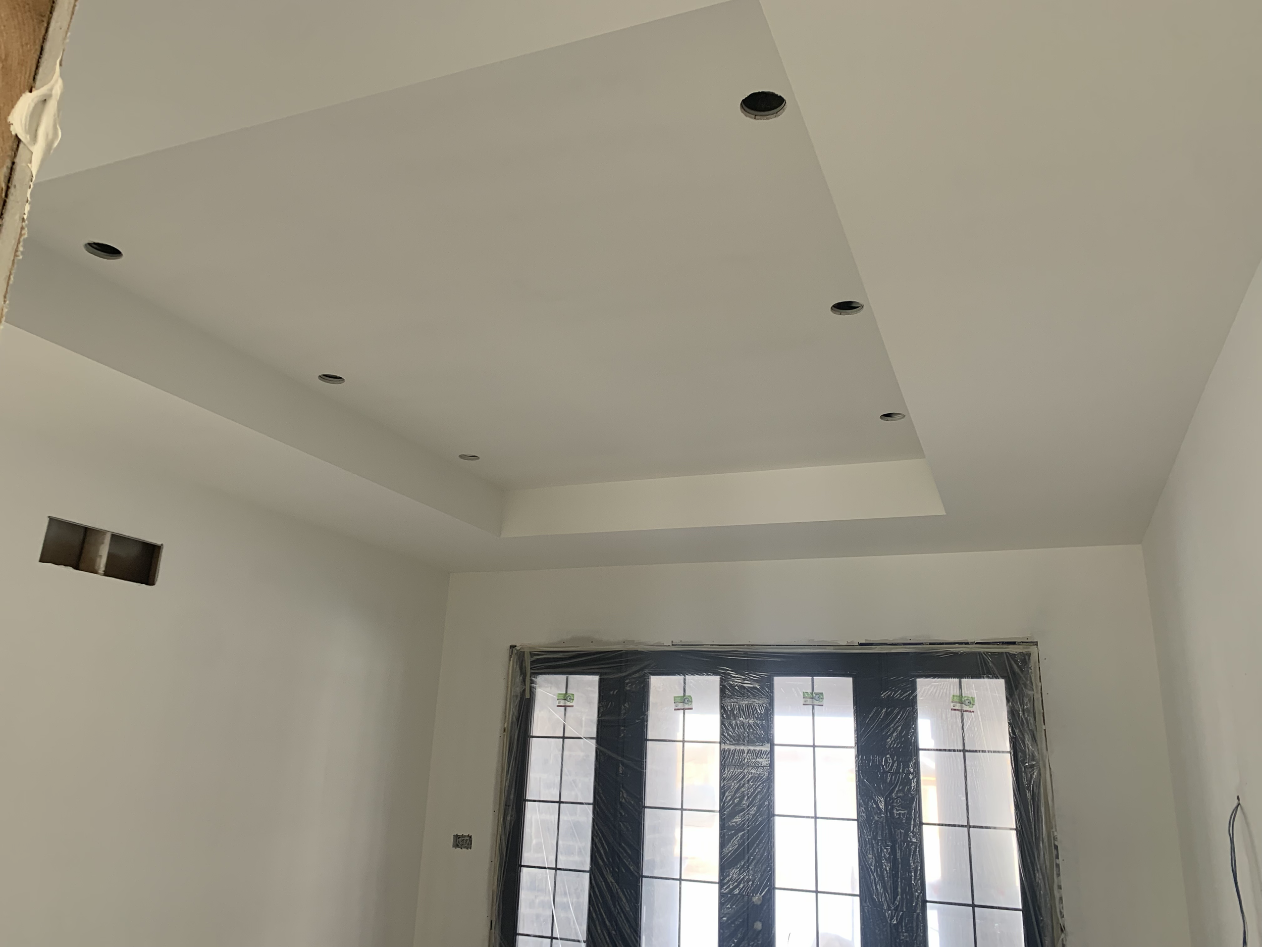 Interior of a room under construction with a tray ceiling and recessed lighting fixtures. There is a large window with black protective film and masking tape around the edges, and the walls are painted white.