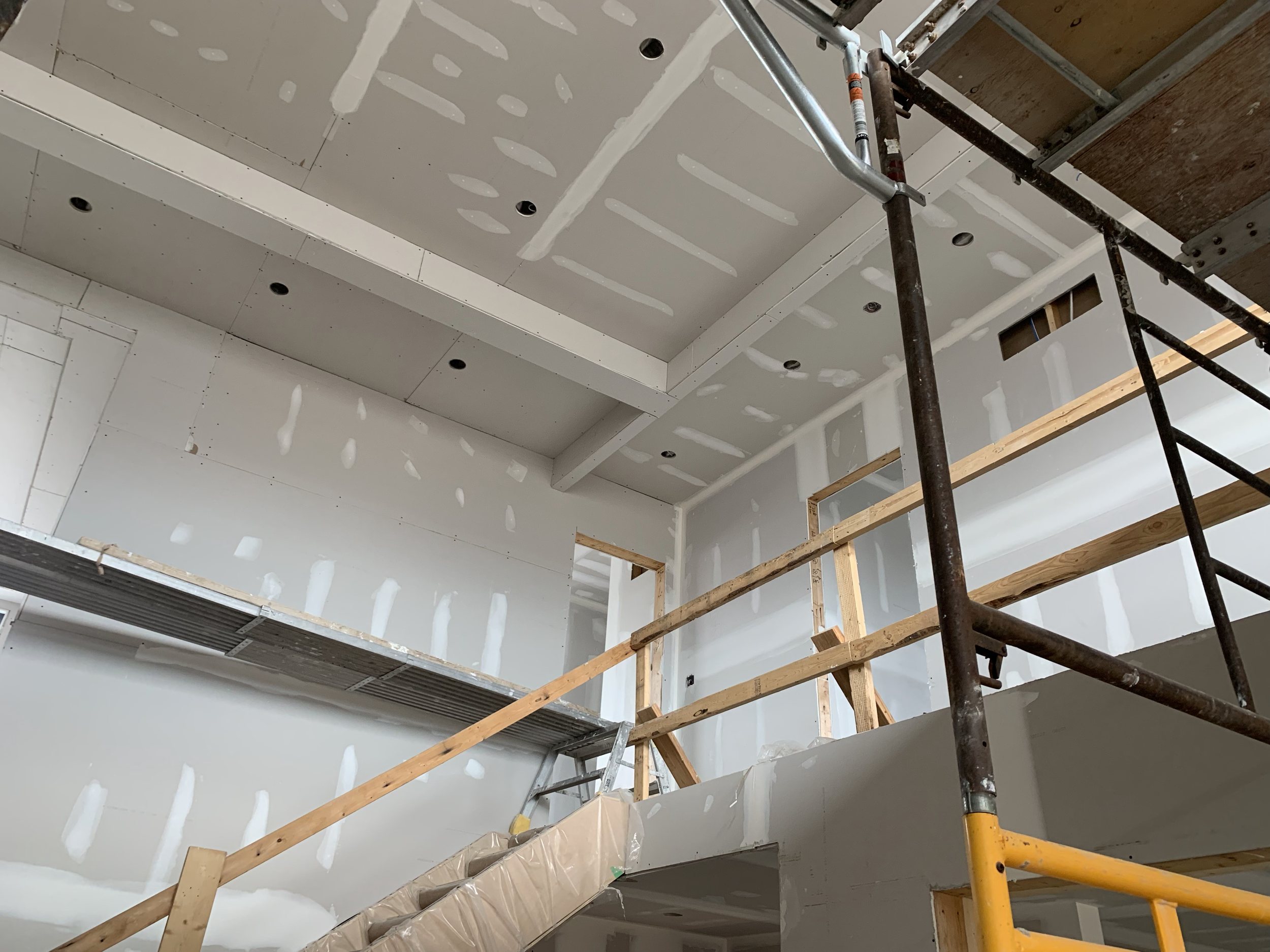 Construction site with drywall being installed on the ceiling and walls. Wooden scaffolding and ladders are present for workers to access higher areas. The drywall is partially finished with joint compound visible at seams.