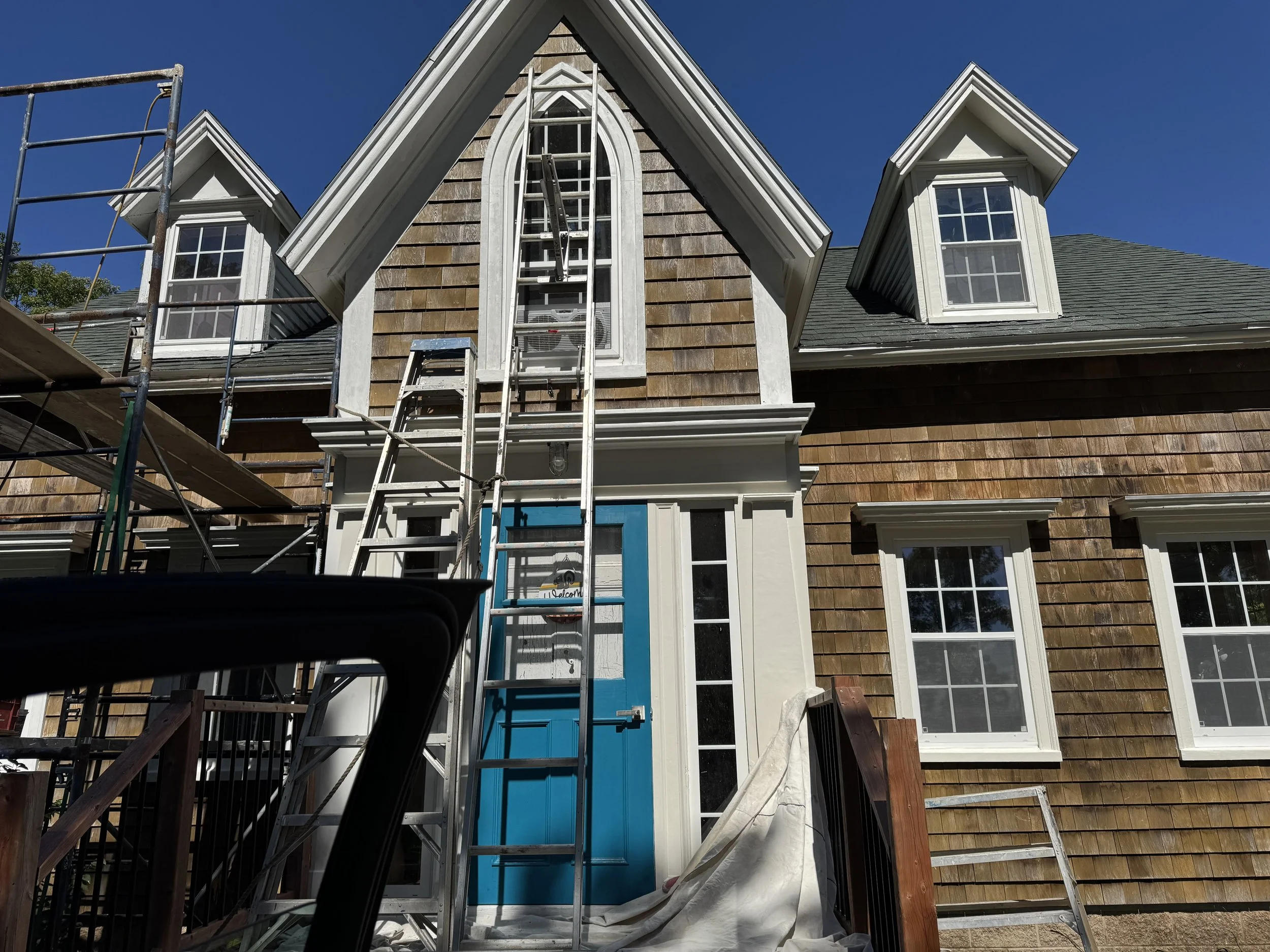 House under construction with a blue front door, scaffolding on the side, and two dormer windows on the roof against a clear blue sky.