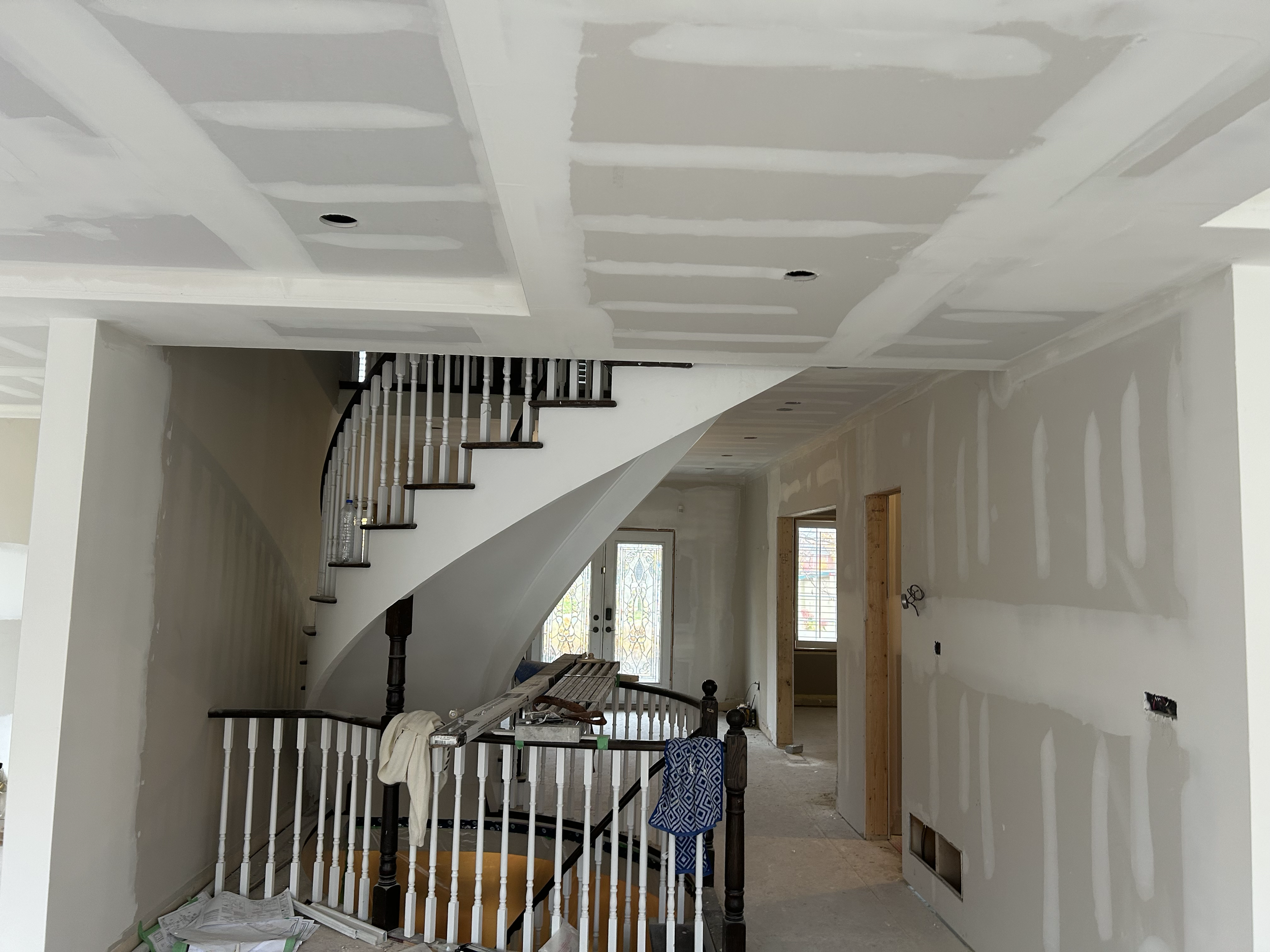 Interior of a house under renovation with drywall installation in progress, staircase with painted white railing, construction materials and tools scattered on the floor.