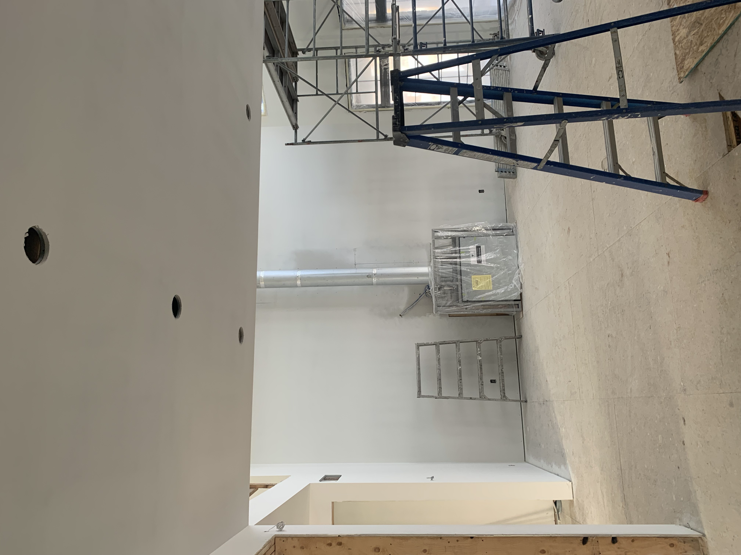 An interior construction site with ladders, scaffolding, and a plastic-covered appliance or piece of equipment, with white walls and a vent pipe.