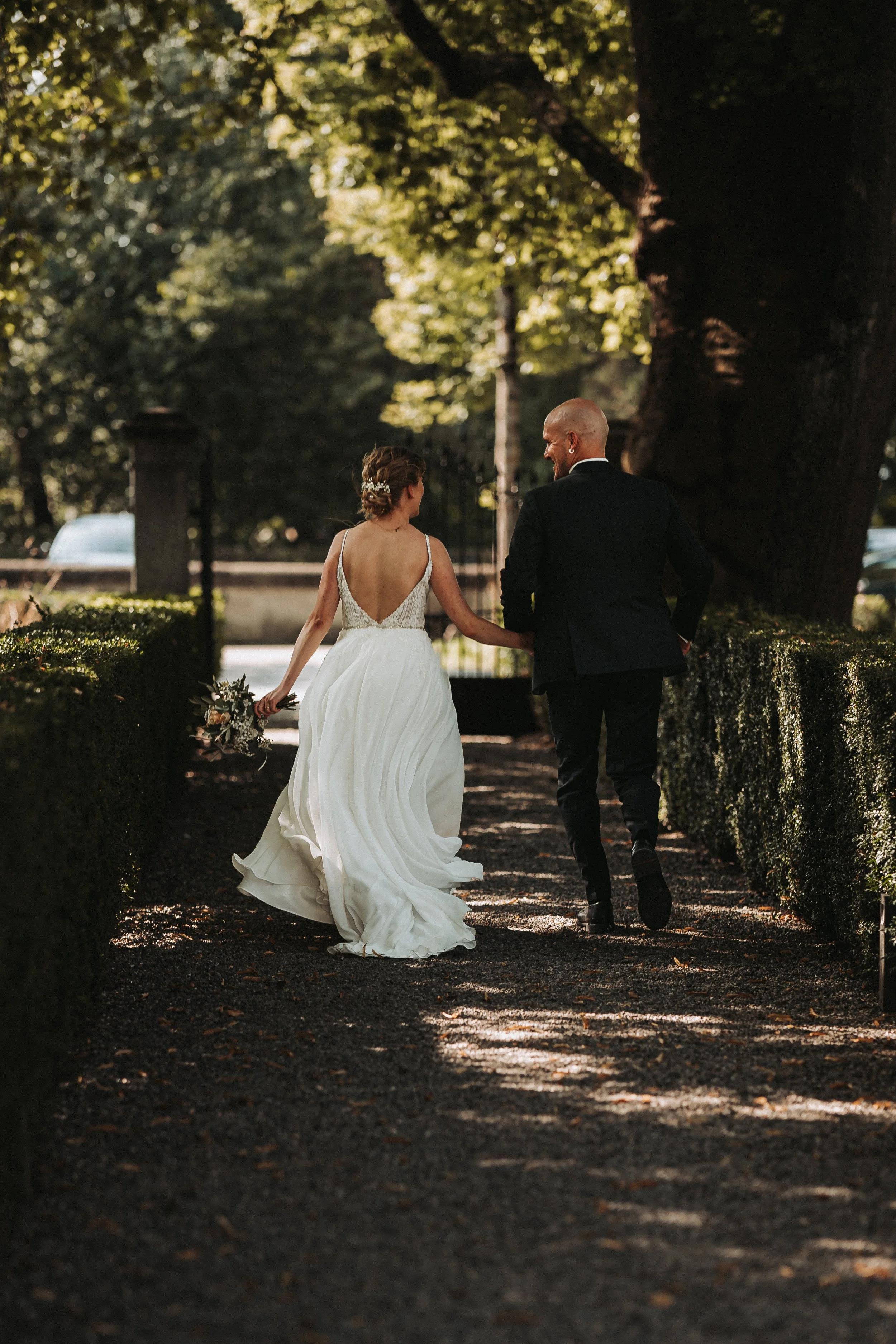 Ein Brautpaar läuft Hand in Hand durch einen schattigen Weg in einem Park, umgeben von Bäumen, bei Sonnenlicht, die Frau trägt ein weißes Hochzeitskleid und hält einen Blumenstrauß, der Mann trägt einen schwarzen Anzug.