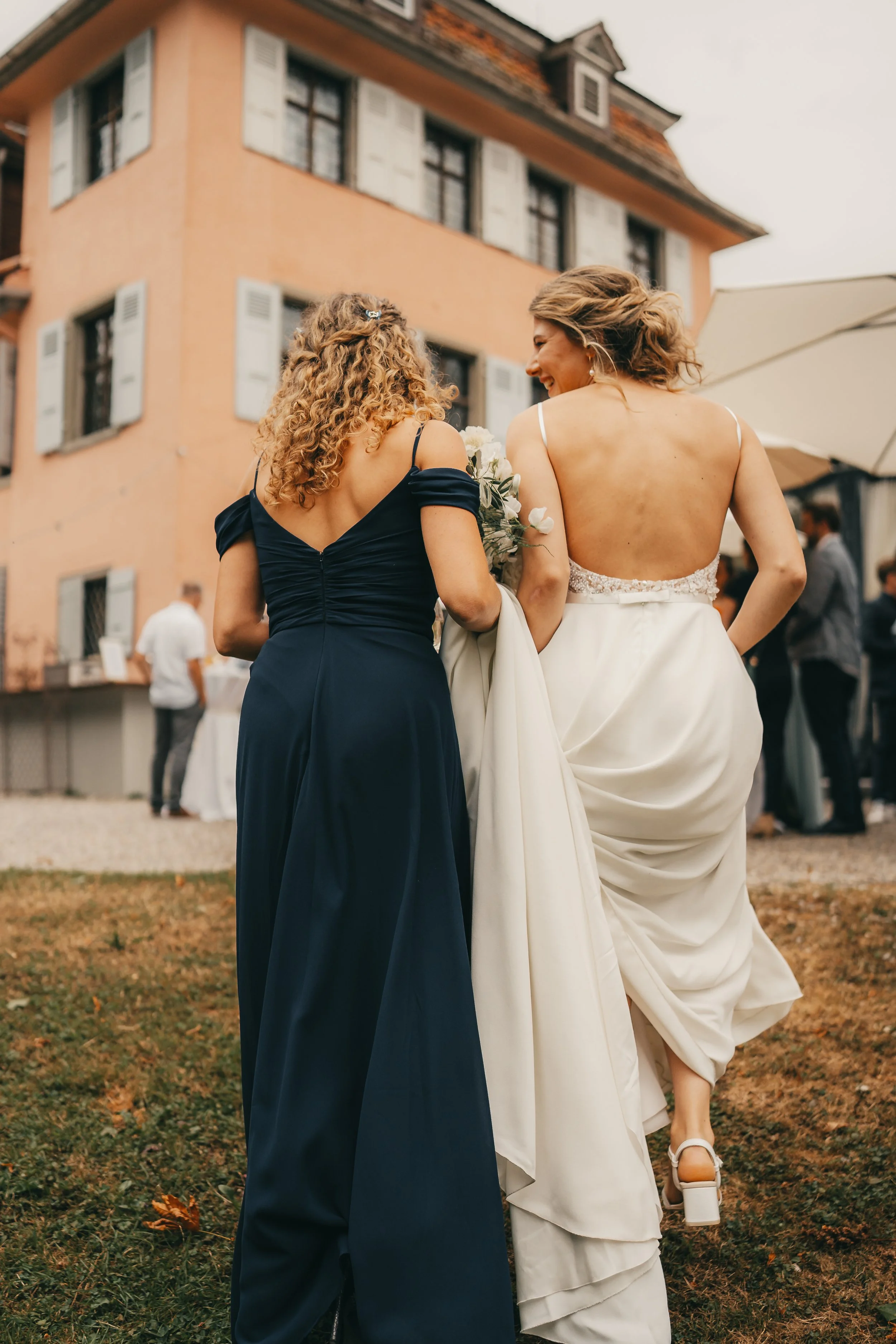 Zwei Frauen bei einer Hochzeitsfeier im Freien, eine trägt ein weißes Brautkleid und die andere ein dunkelblaues Kleid, lachen und unterhalten sich, im Hintergrund ein mehrstöckiges Haus mit offenen Fenstern, Gäste und Dekorationen