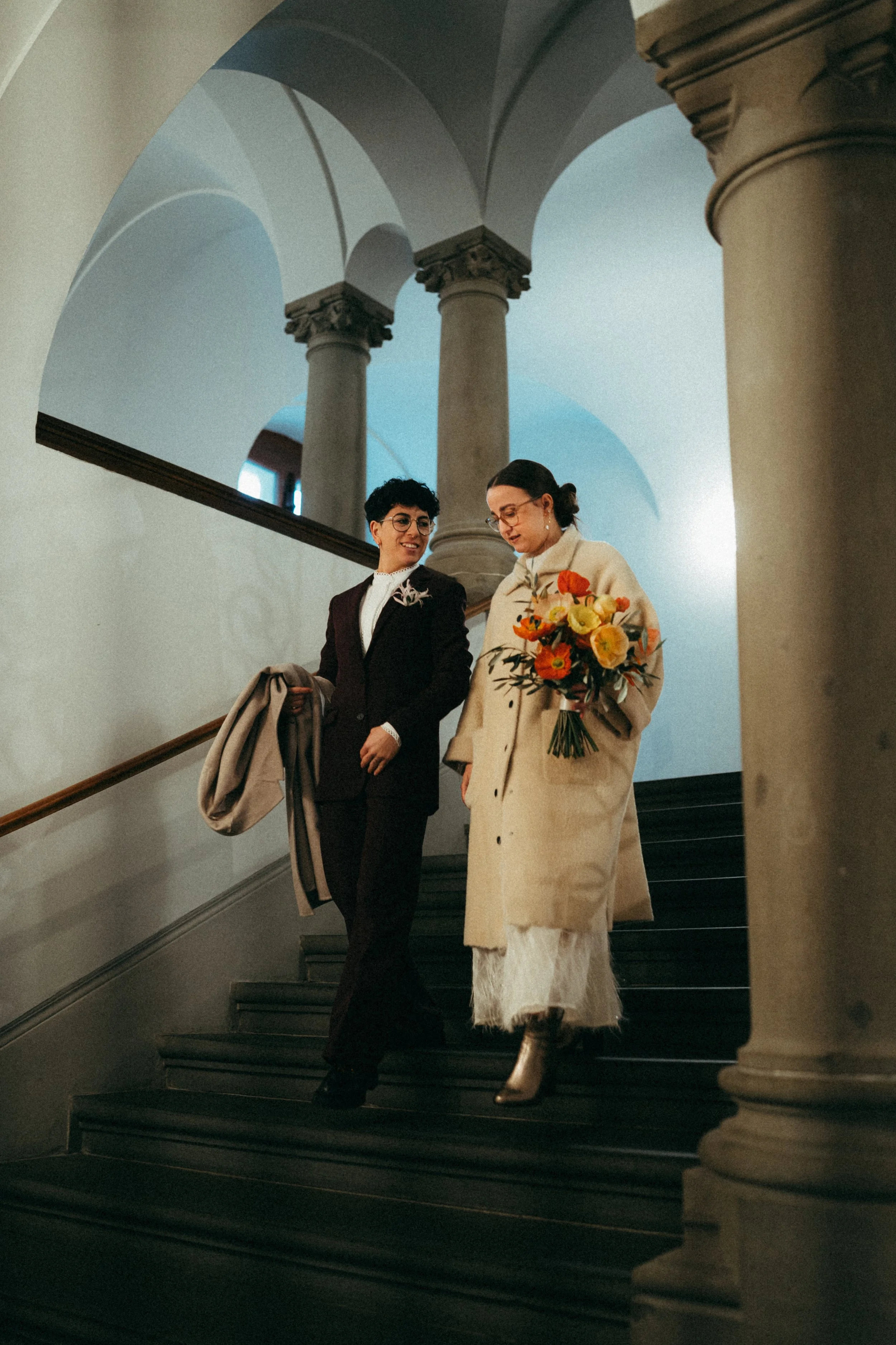 Zwei Frauen gehen die Treppe in einem historischen Gebäude hinunter. Eine hält einen Blumenstrauß, die andere trägt einen Mantel und hat eine Tasche in der Hand.