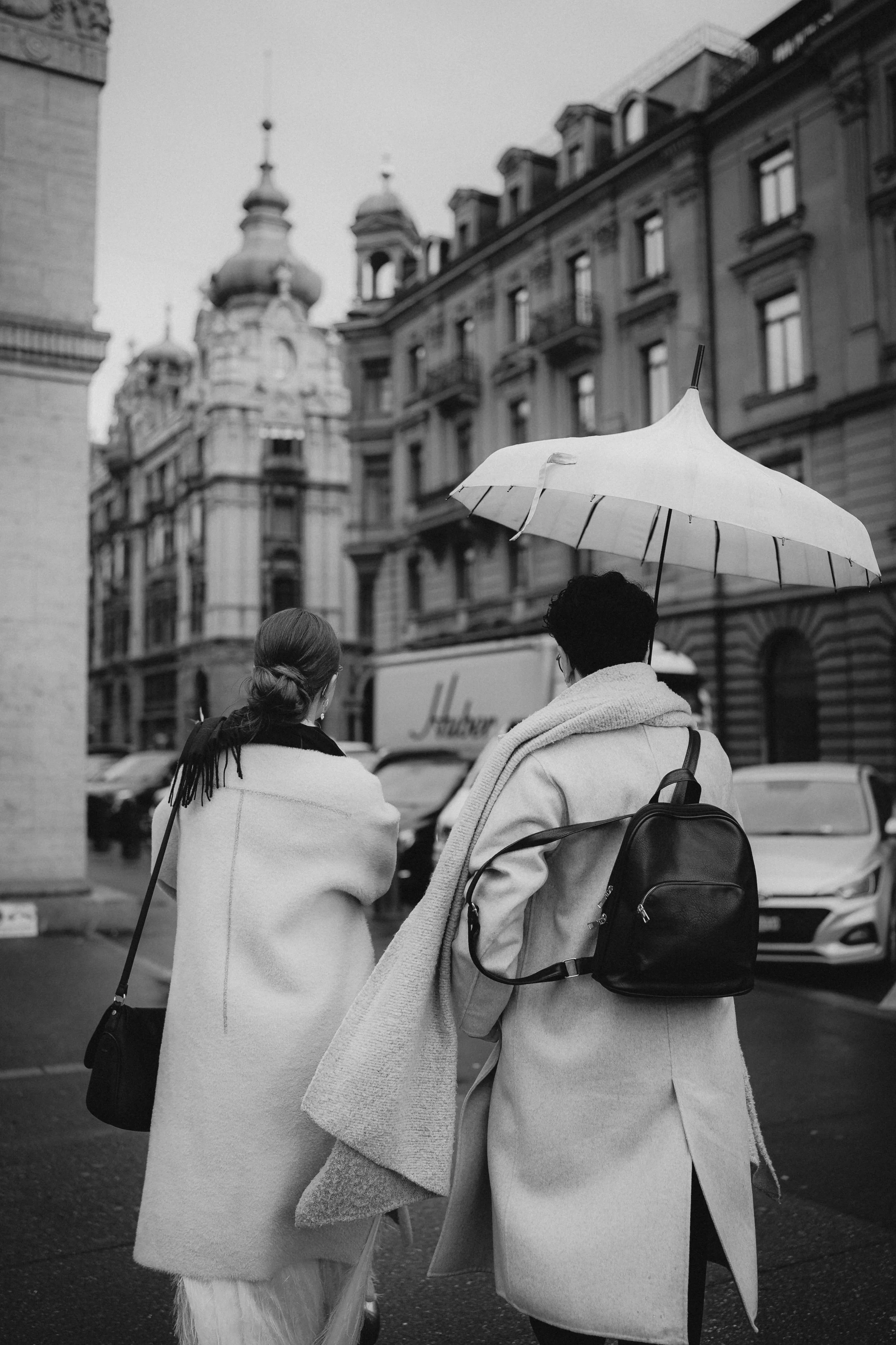 Zwei Frauen stehen auf einer Stadtstraße bei Regen, eine hält einen weißen Regenschirm und trägt einen weißen Mantel, die andere trägt ebenfalls einen Mantel. Im Hintergrund sind historische Gebäude und Autos zu sehen.