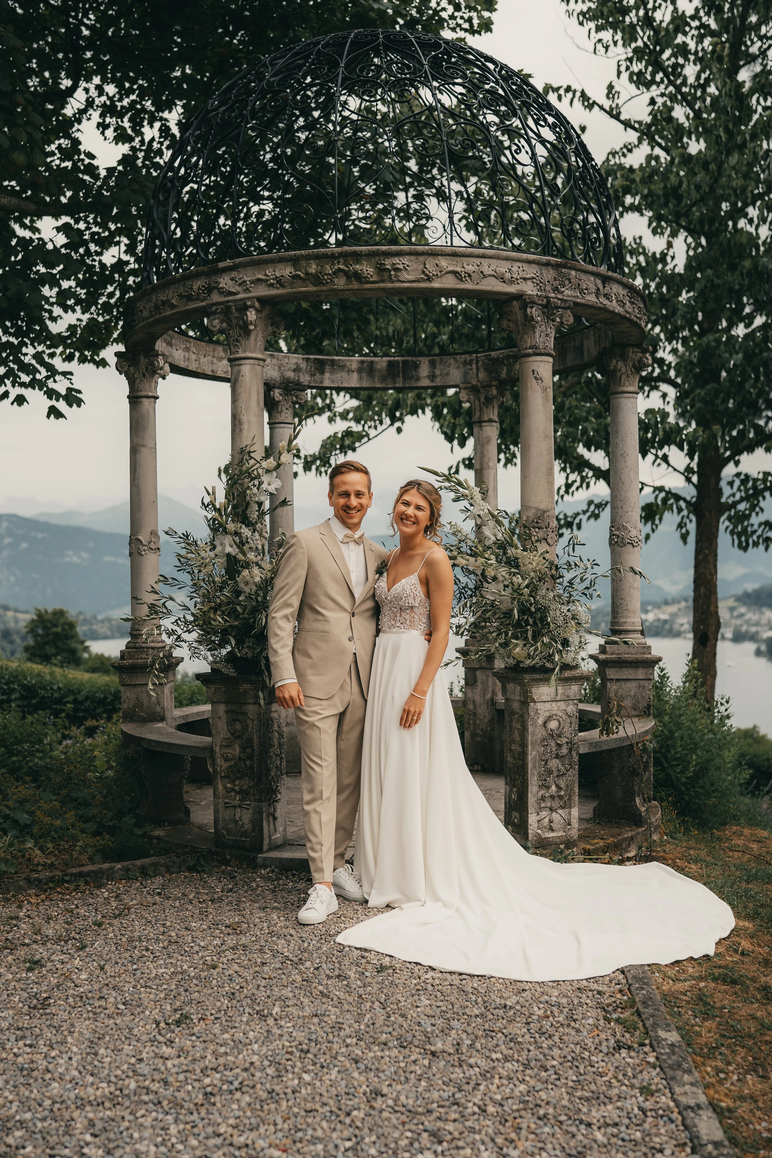 Ein glückliches Brautpaar, stehende vorne in einem weißem Brautkleid und einem beigen Anzug, vor einem romantischen Pavillon mit grünem Baum- und Bergkulisse im Hintergrund.