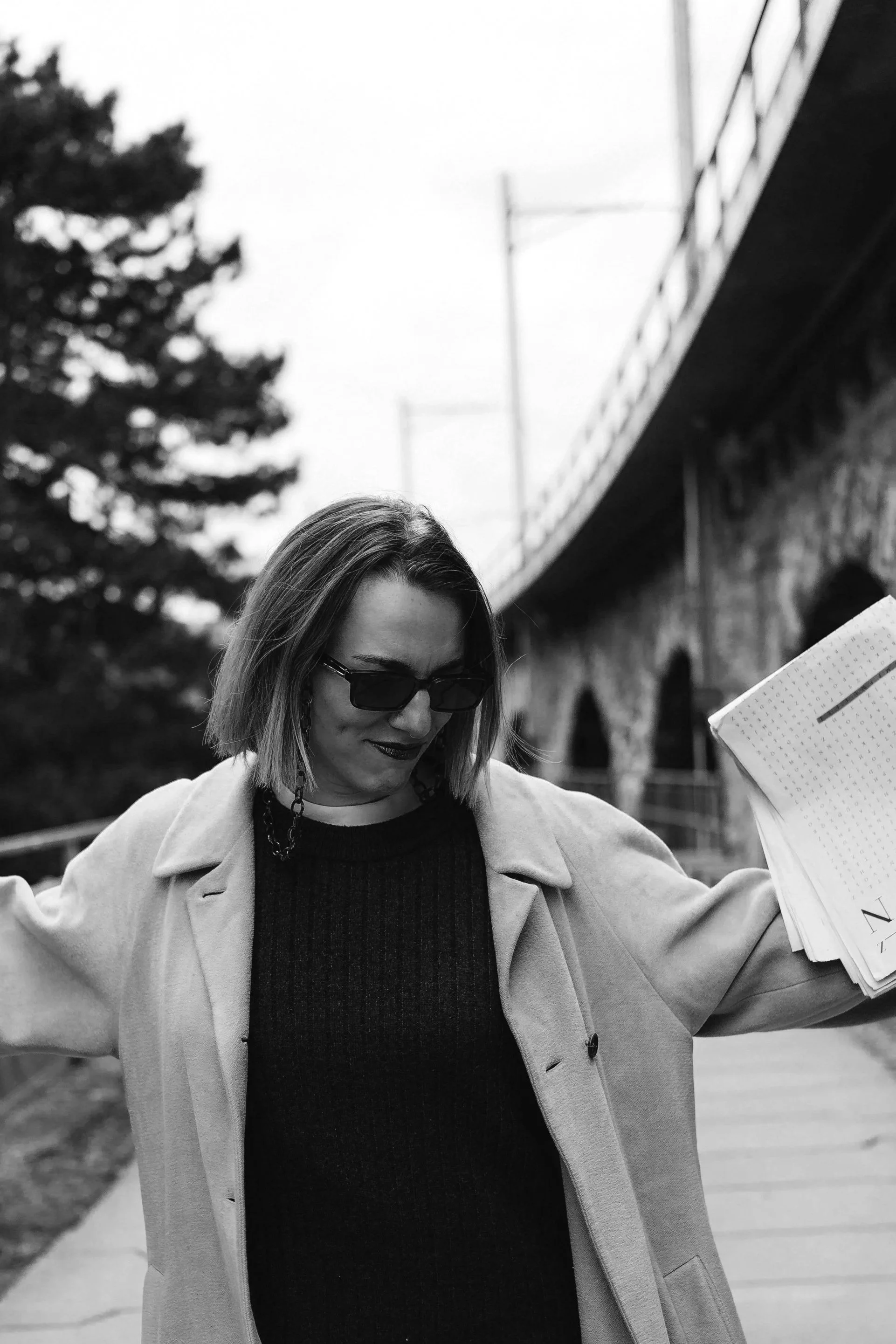 Schwarz-weiß-Fotografie einer Frau mit long bob, Sonnenbrille, schwarzem Kleid und heller Jacke, die in einer urbanen Umgebung steht.