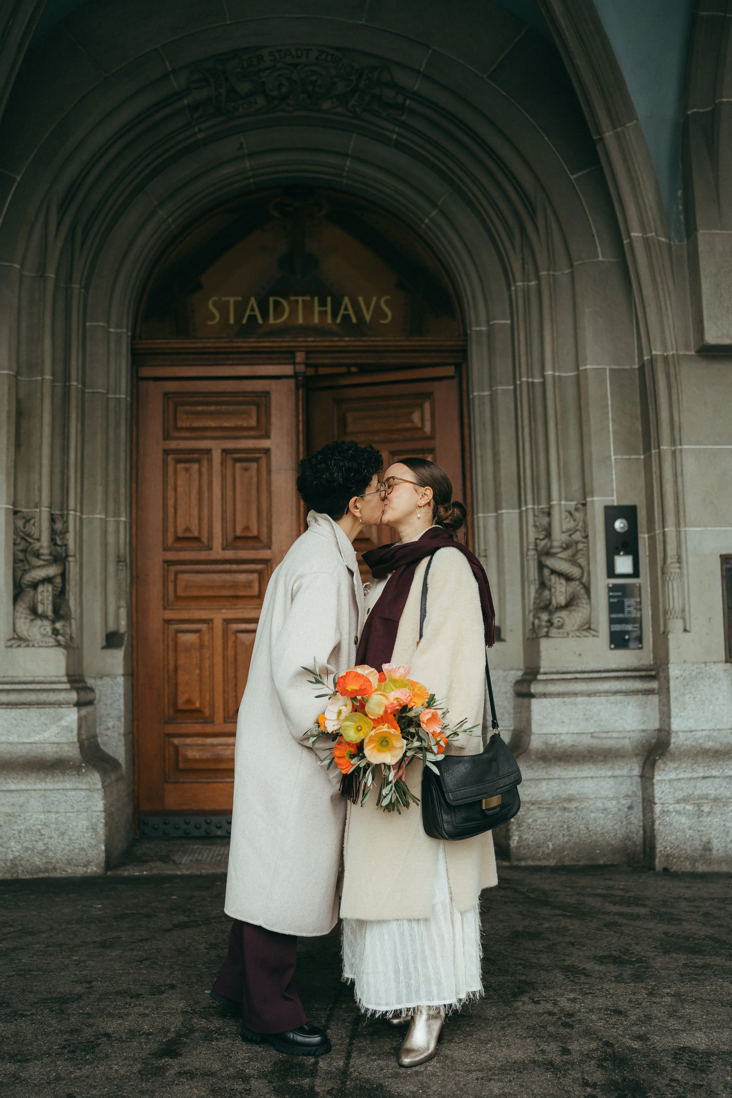 Zwei Frauen küssen sich vor einem historischen Gebäudetor mit dem Schriftzug "Stadthaus". Eine Frau hält einen Blumenstrauß. Sie tragen elegante Kleidung.
