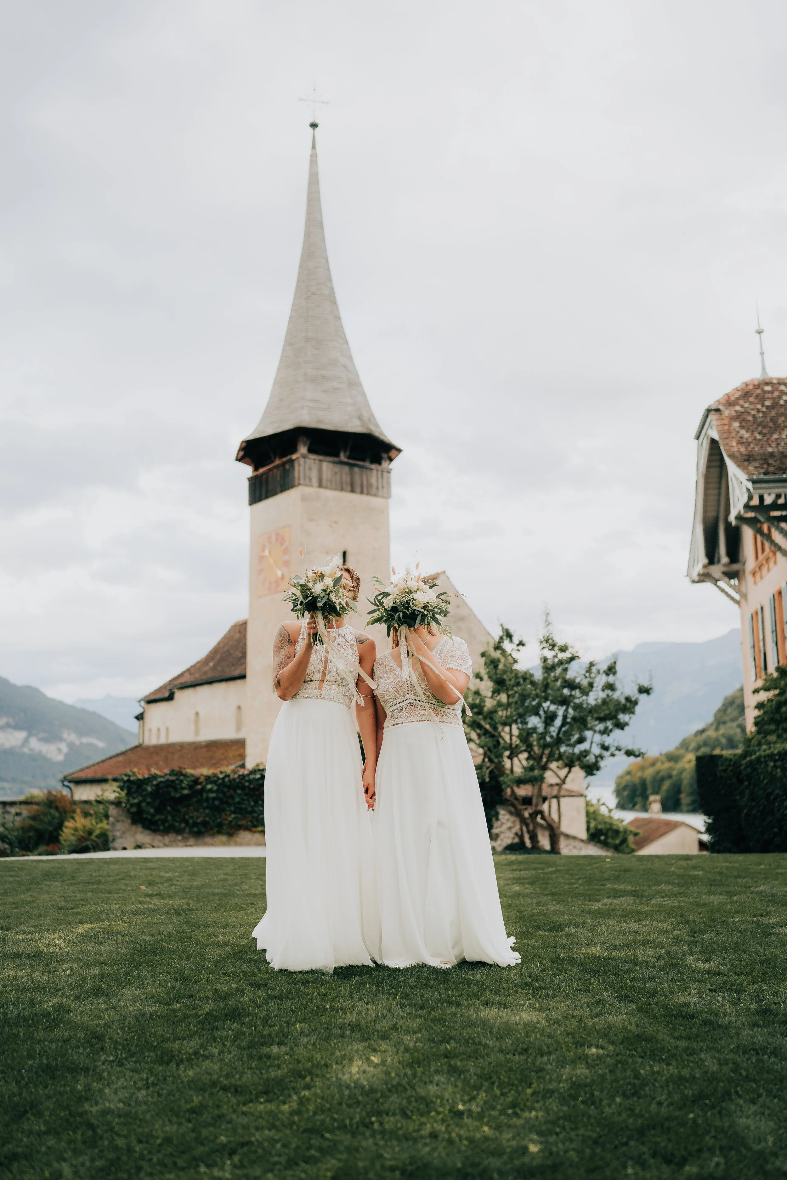 Zwei Frauen in weißen Brautkleidern stehen nebeneinander auf einem Rasen vor einer Kirche, ihre Gesichter sind von großen Blumensträußen verdeckt. Der Himmel ist bewölkt, im Hintergrund sind Berge und traditionelle Gebäude sichtbar.