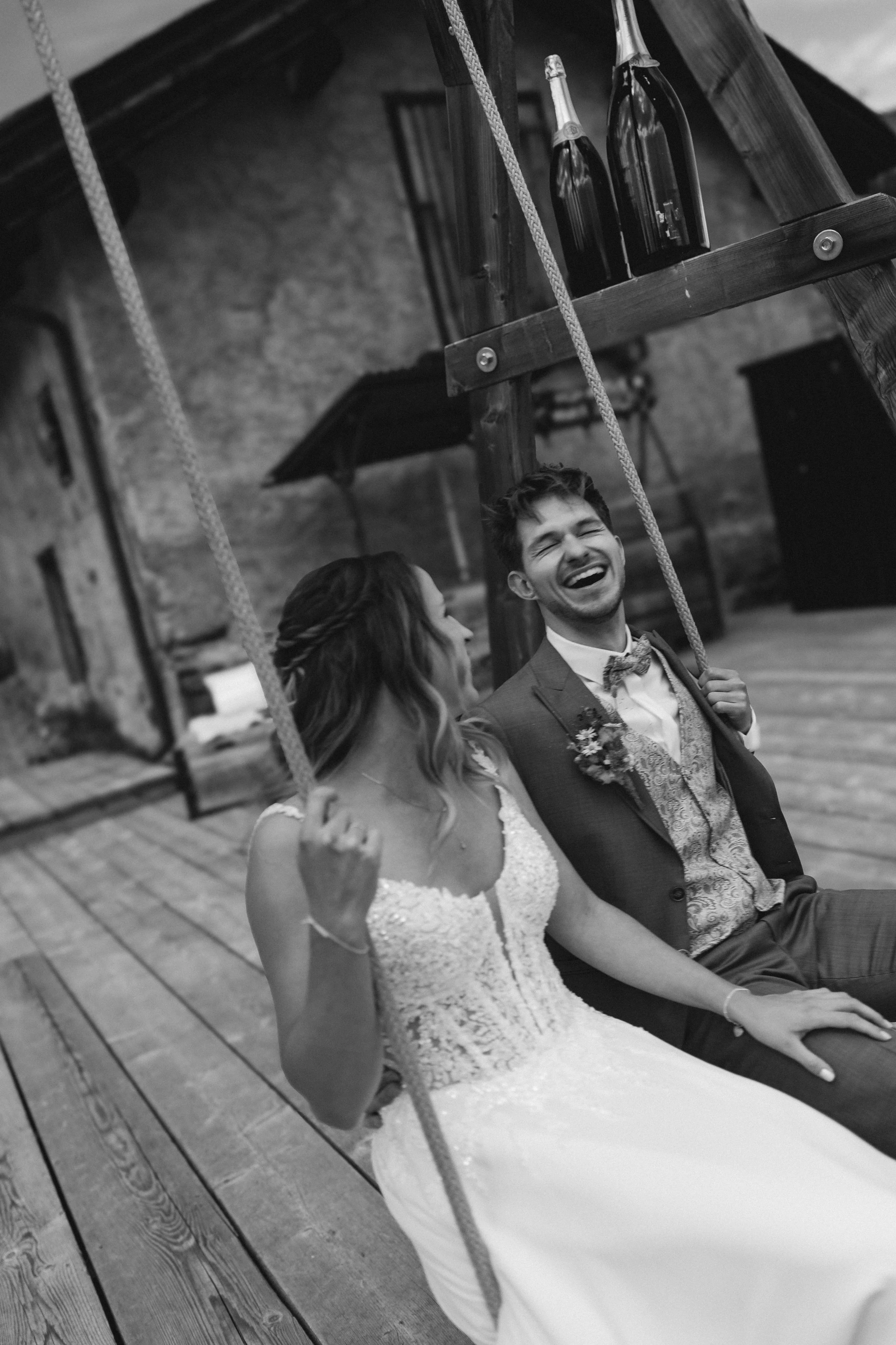 Ein lächelnder Mann und eine Frau auf einer Schaukel im Freien, im Hintergrund eine Steinfassade und ein Regenschirm. Die Frau trägt ein weißes Hochzeitskleid, der Mann einen Anzug mit Fliege.