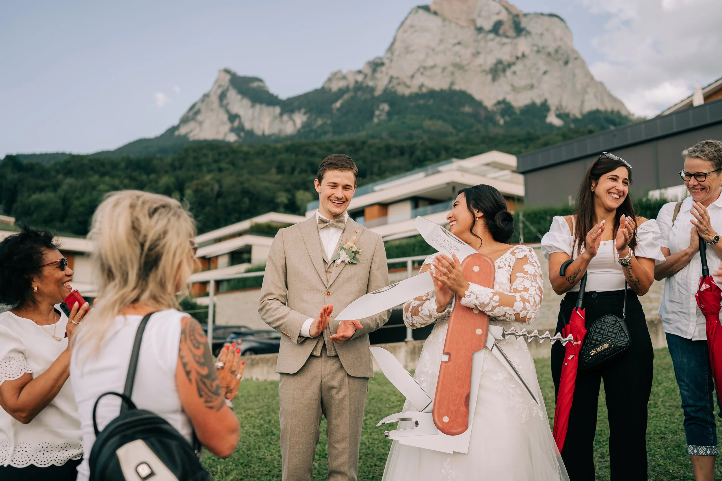 Menschen bei einer Hochzeitsfeier im Freien, im Hintergrund Berge und modernes Gebäude, die Braut und der Bräutigam umgeben von Freunden.