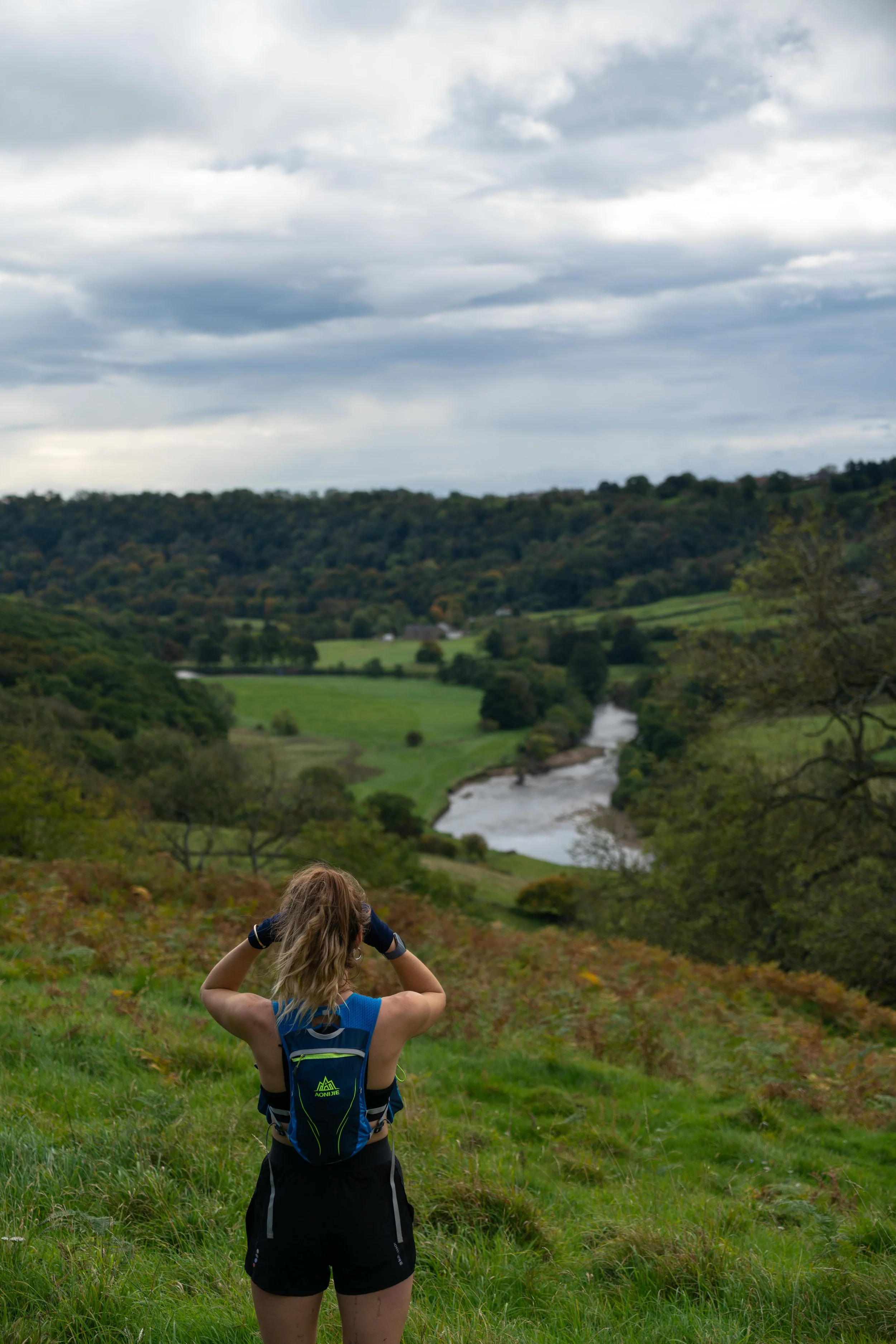 A woman standing on a grassy hill with a river and green hills in the background, under a cloudy sky.