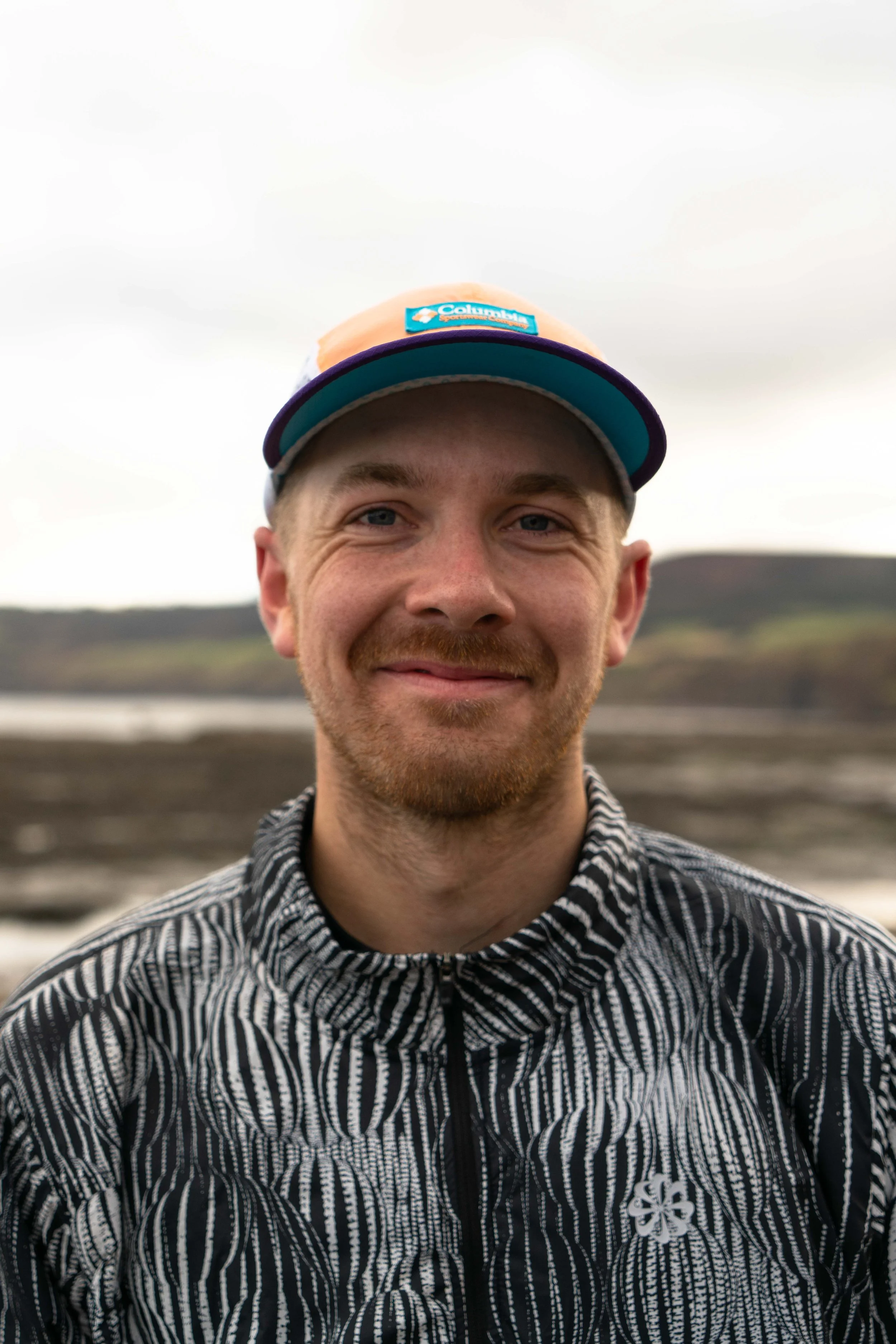 A smiling man with blue eyes and facial hair wearing a black and white patterned jacket and a multicolored Columbia hat, outdoors with a blurred natural landscape in the background.