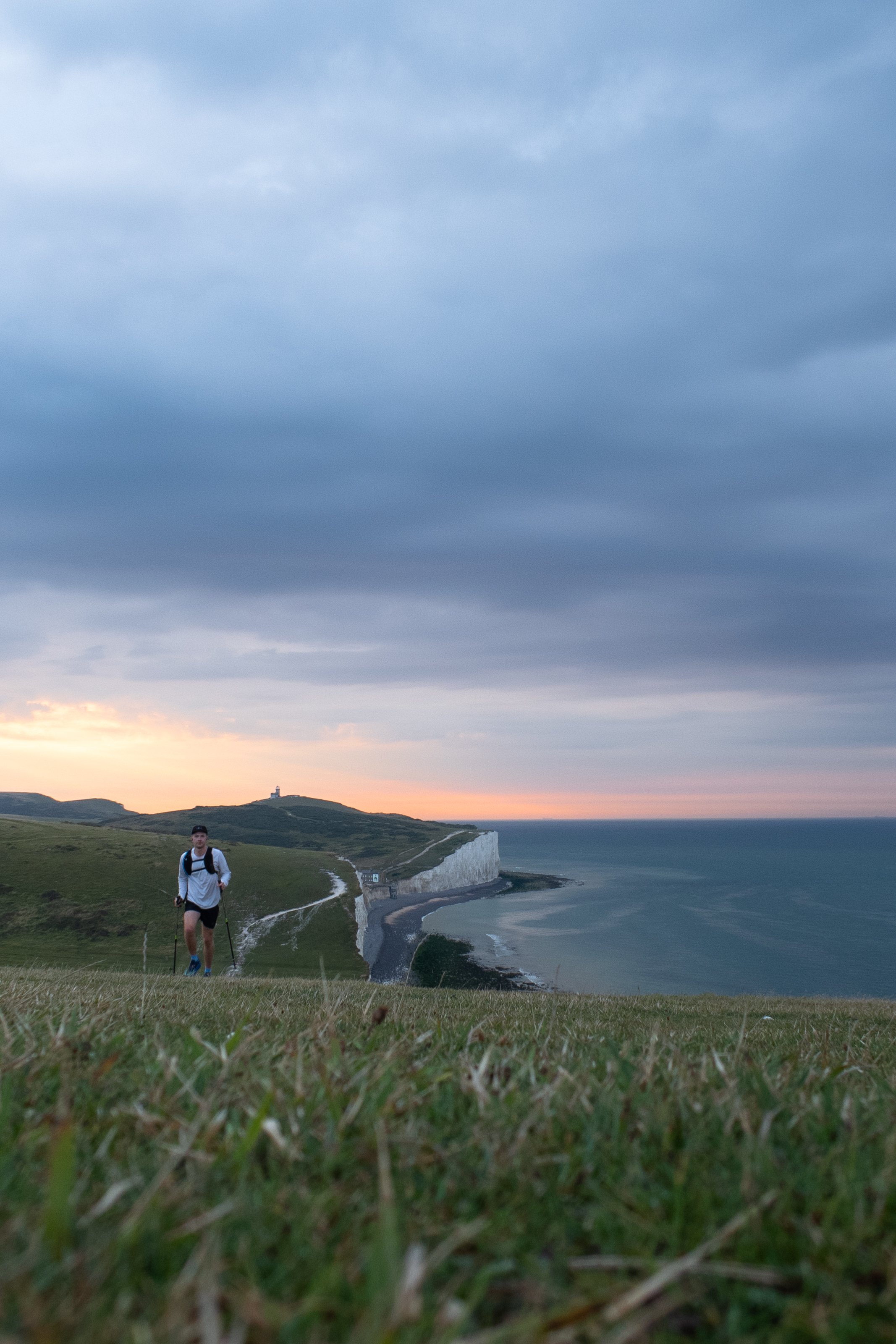 A person hiking along a grassy cliffside trail overlooking the ocean at sunset, with white cliffs and a lighthouse in the distance.