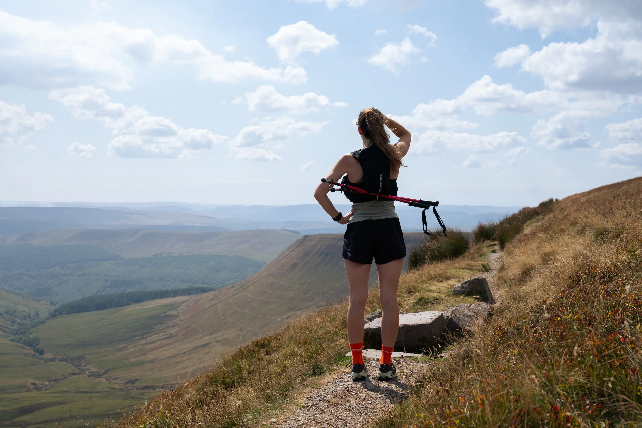 A woman with long hair, wearing a black sleeveless top, black shorts, red socks, and hiking shoes, standing on a trail on a hillside looking out over a panoramic view of rolling green hills and valleys under a partly cloudy sky, holding a hiking pole over her shoulders.