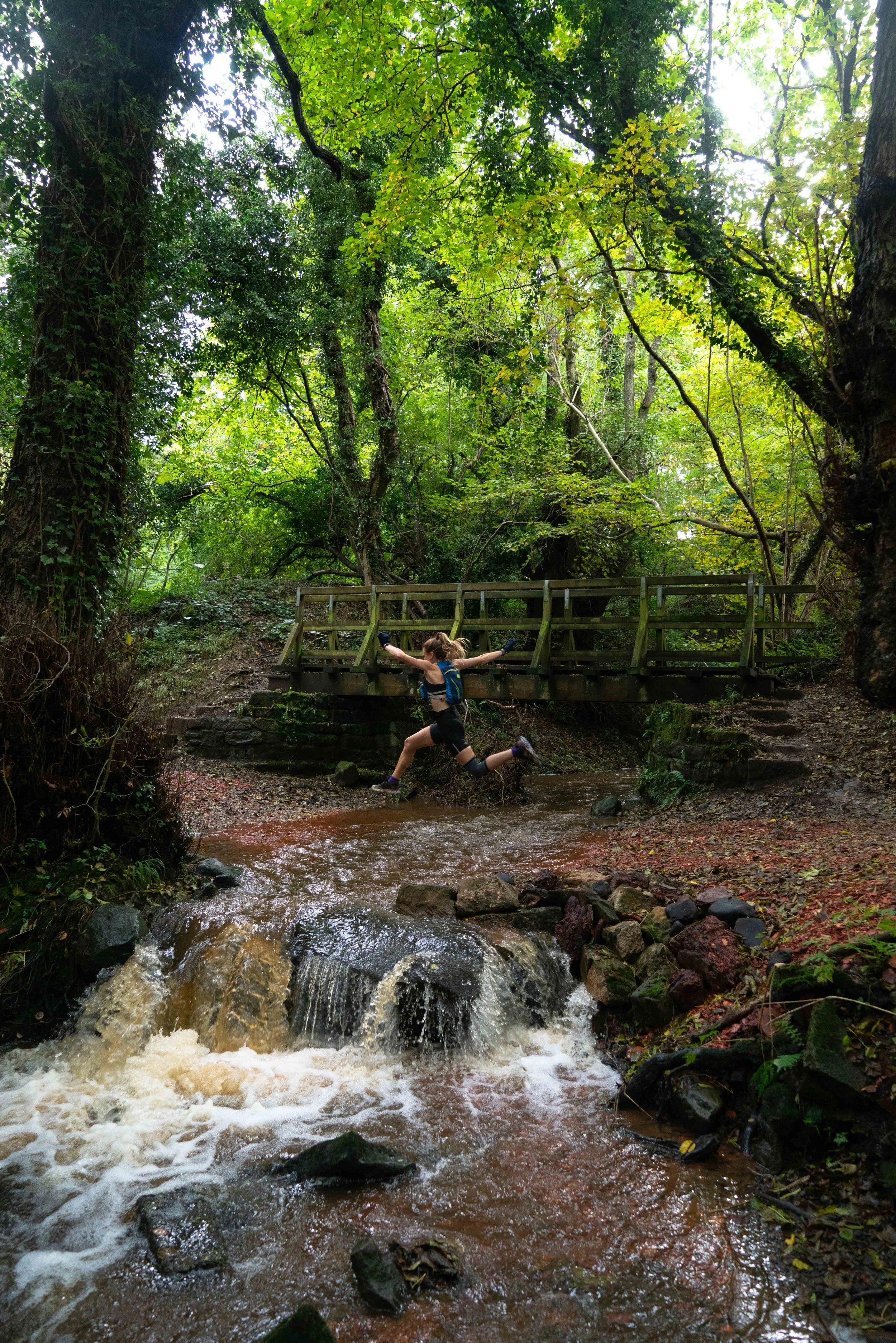 A person in a blue tank top and black shorts jumping over a small stream in a lush green forest. The person is wearing a backpack and black gloves, with arms outstretched and one leg bent mid-air. A wooden bridge is visible in the background, surrounded by tall, leafy trees.