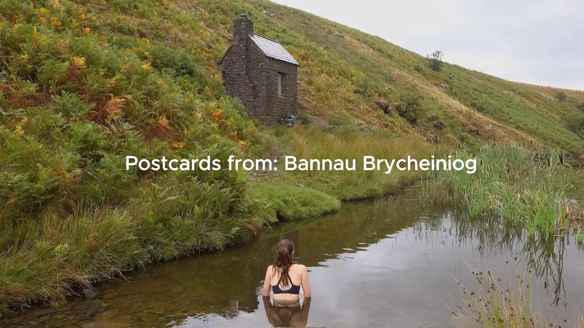 People swimming in a river with a stone house on a hill in the background, surrounded by green and yellow foliage.