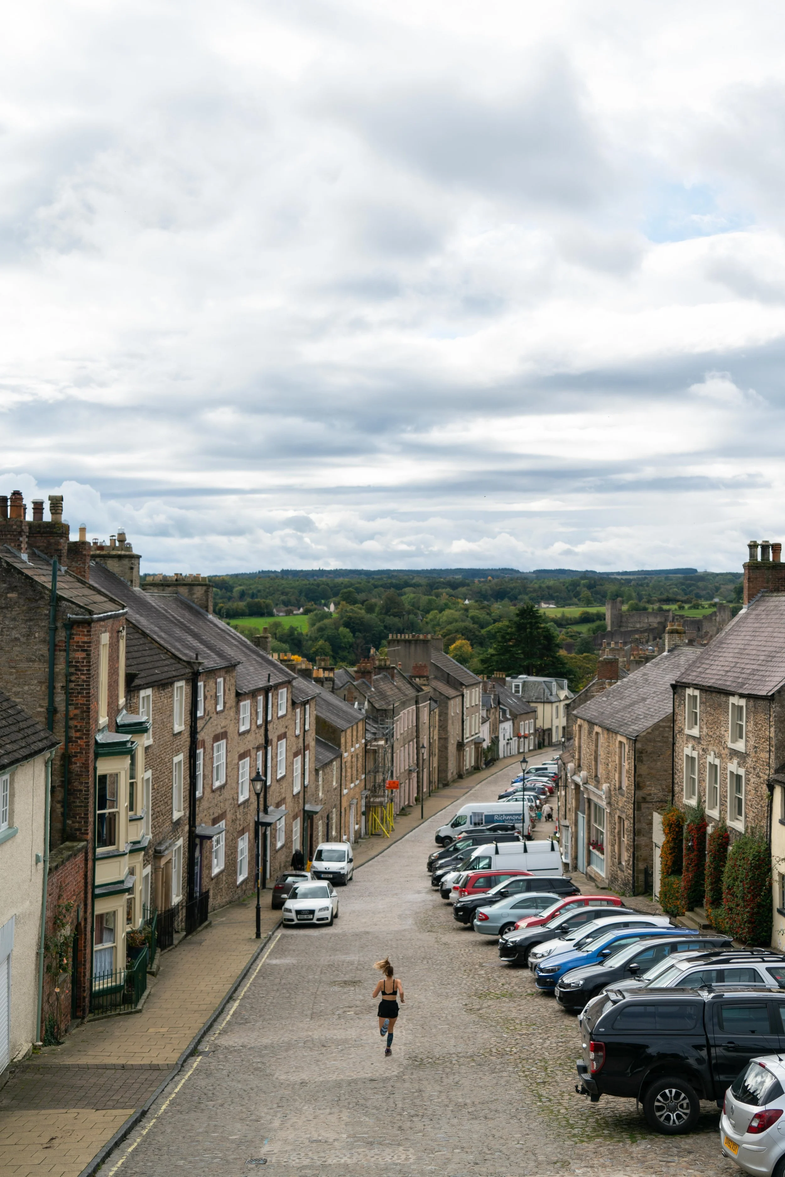 A street view with parked cars lining the side of cobblestone road, old brick buildings on either side, and a woman in athletic wear running down the street with green fields and a cloudy sky in the background.
