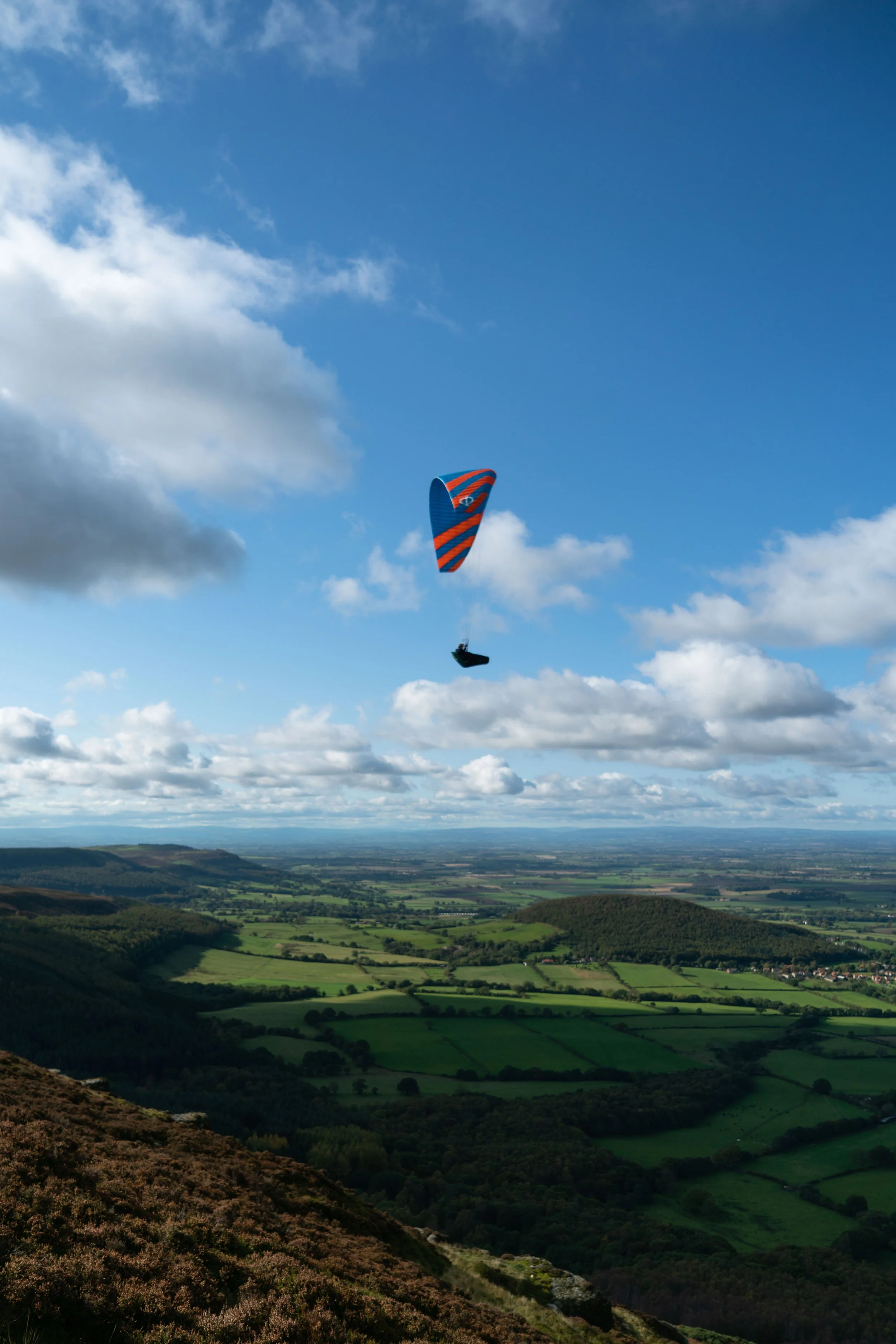 A paraglider with a blue and orange striped wing soaring above a green valley with rolling hills and a partly cloudy sky.