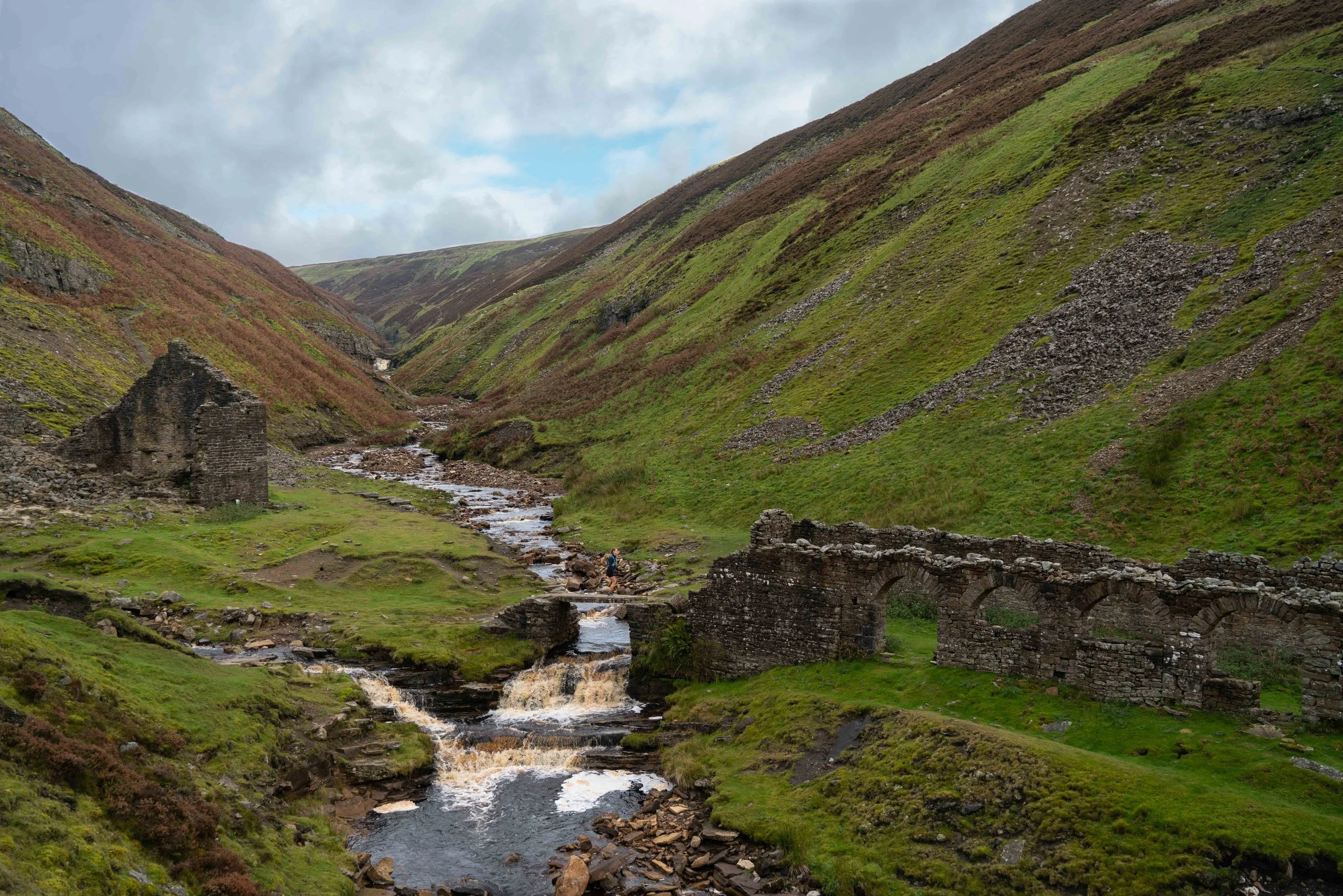 A lush green valley with a small stream running through it, flanked by steep hillsides covered in grass and shrubs. There are ruins of an old stone bridge in the foreground and a person wading in the stream.