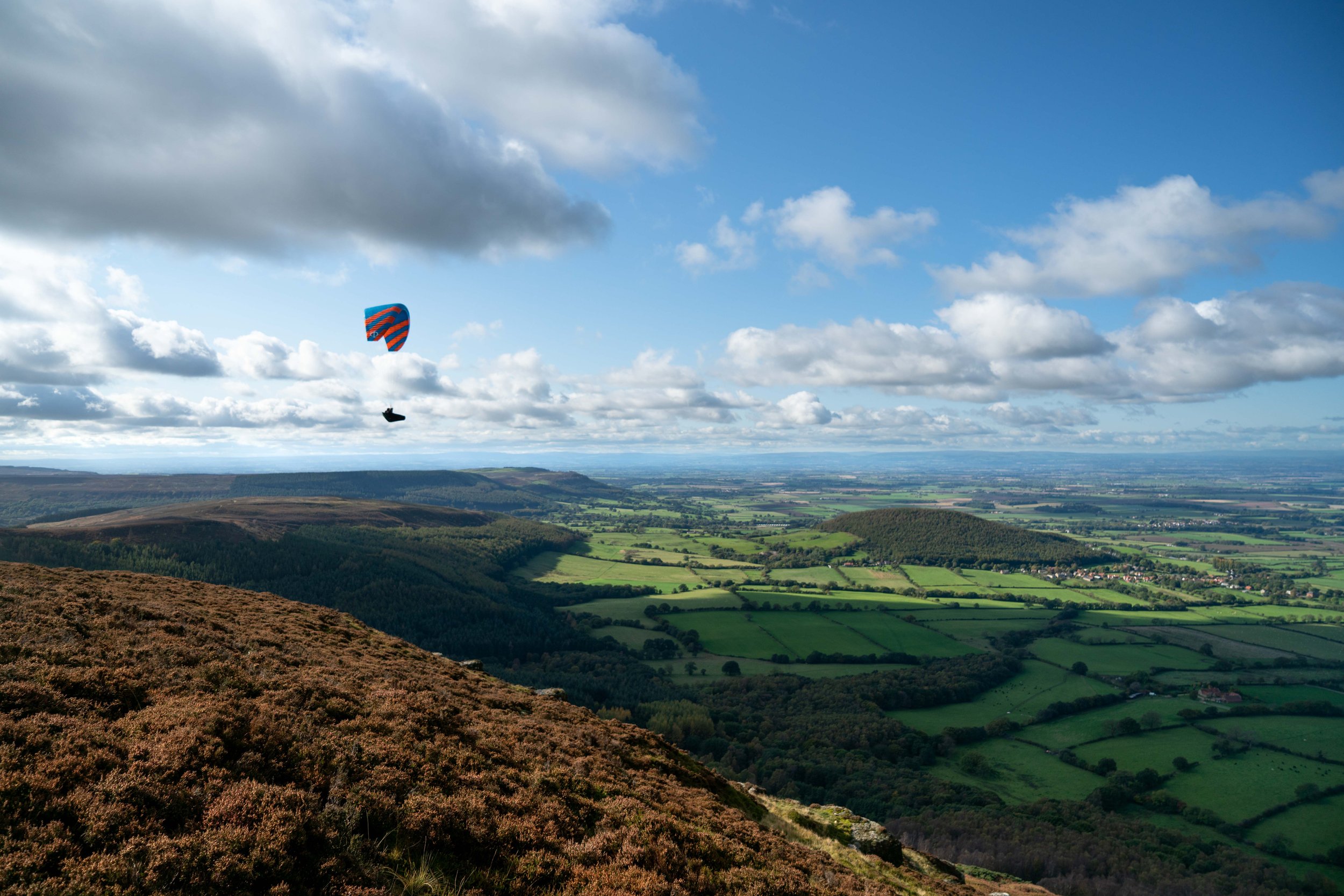 Landscape with green fields, hills, and a cloudy blue sky, featuring a paraglider with a blue and orange striped canopy.