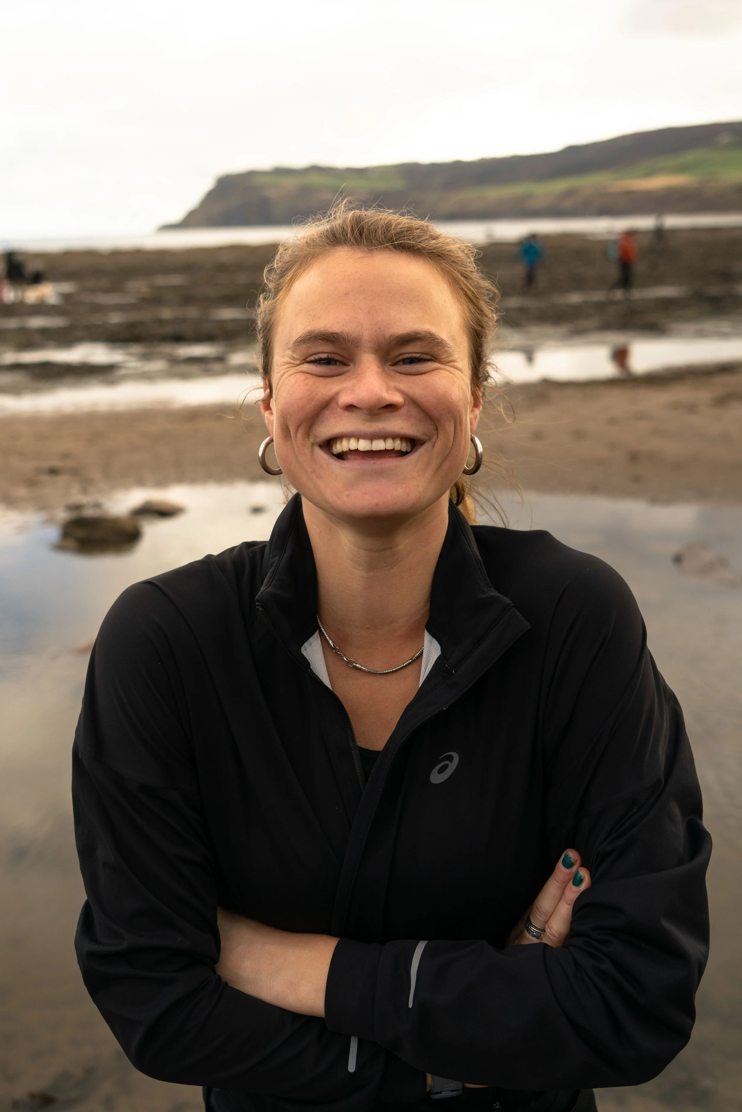 A smiling woman with earrings and necklaces, wearing a black jacket, standing on a beach with a water and cliff background and other people in the distance.