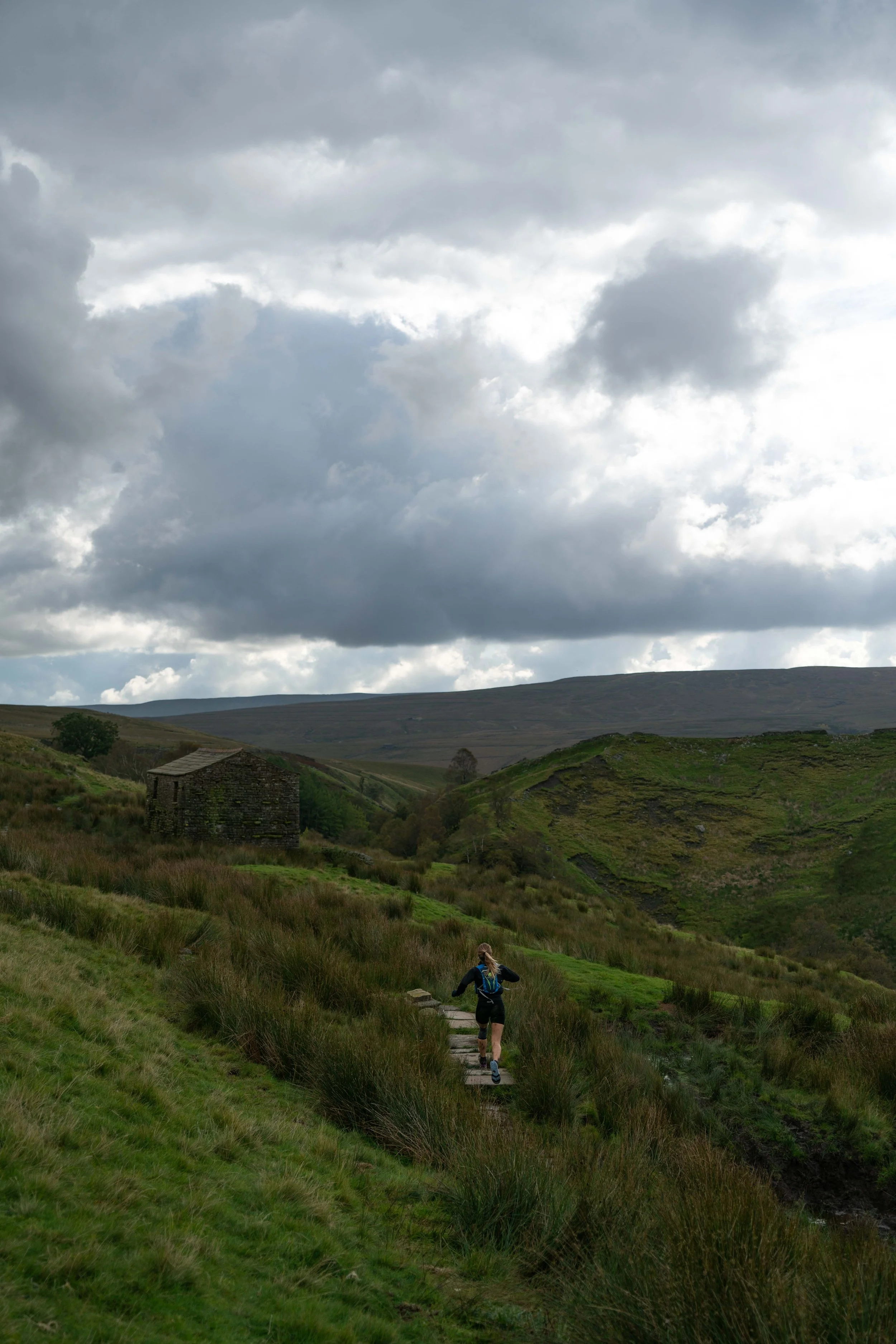 A person hiking on a trail through green rolling hills under a cloudy sky with dark, heavy clouds.