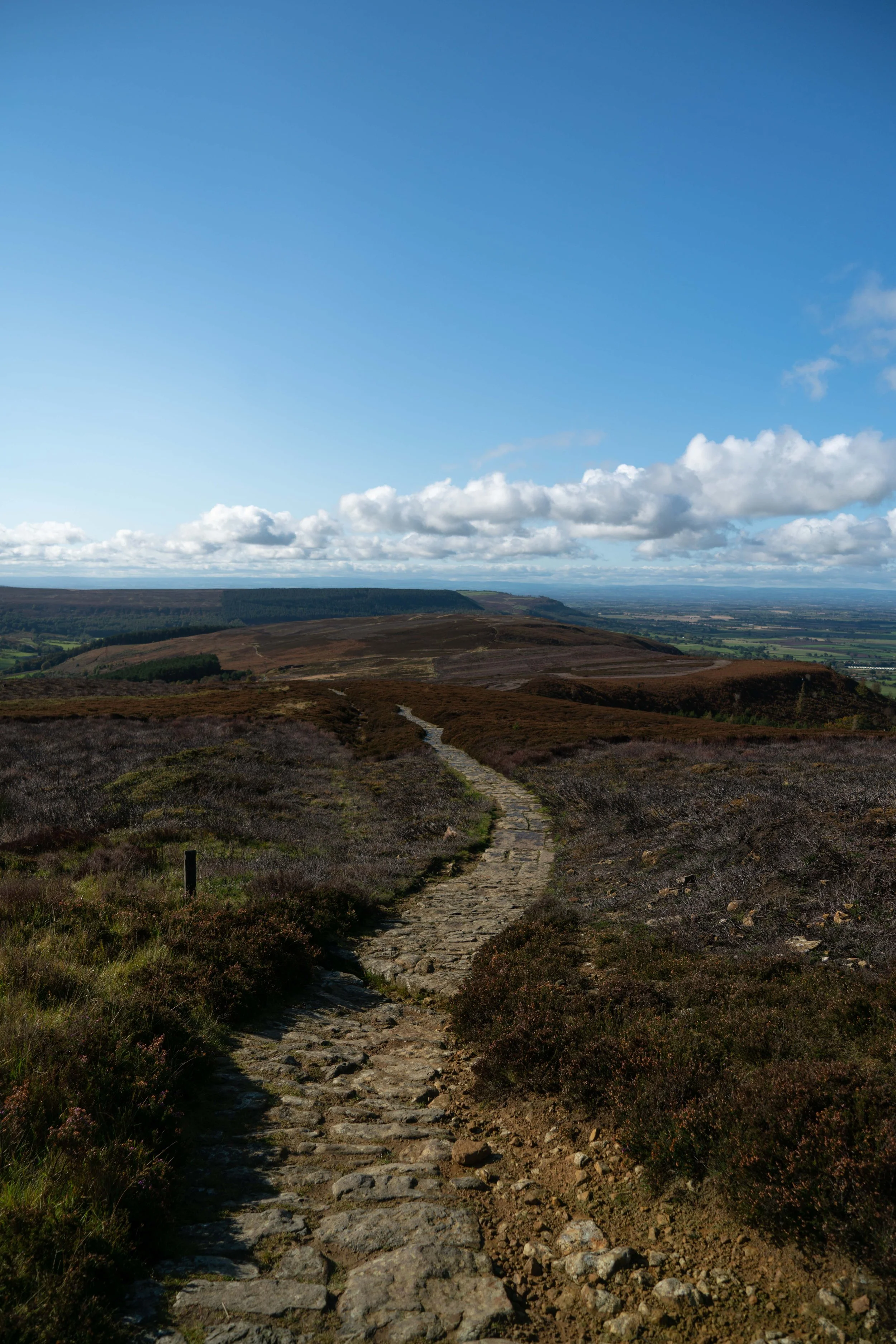 A stone trail leading through a hilly landscape with brown and green vegetation, under a partly cloudy sky with blue sky and white clouds.