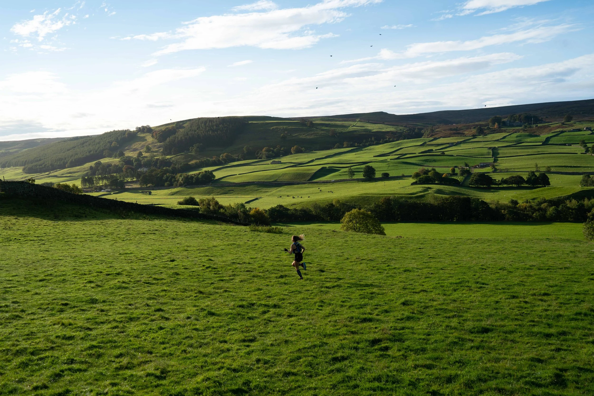 A person running on a lush green field with rolling hills and a partly cloudy sky in the background.