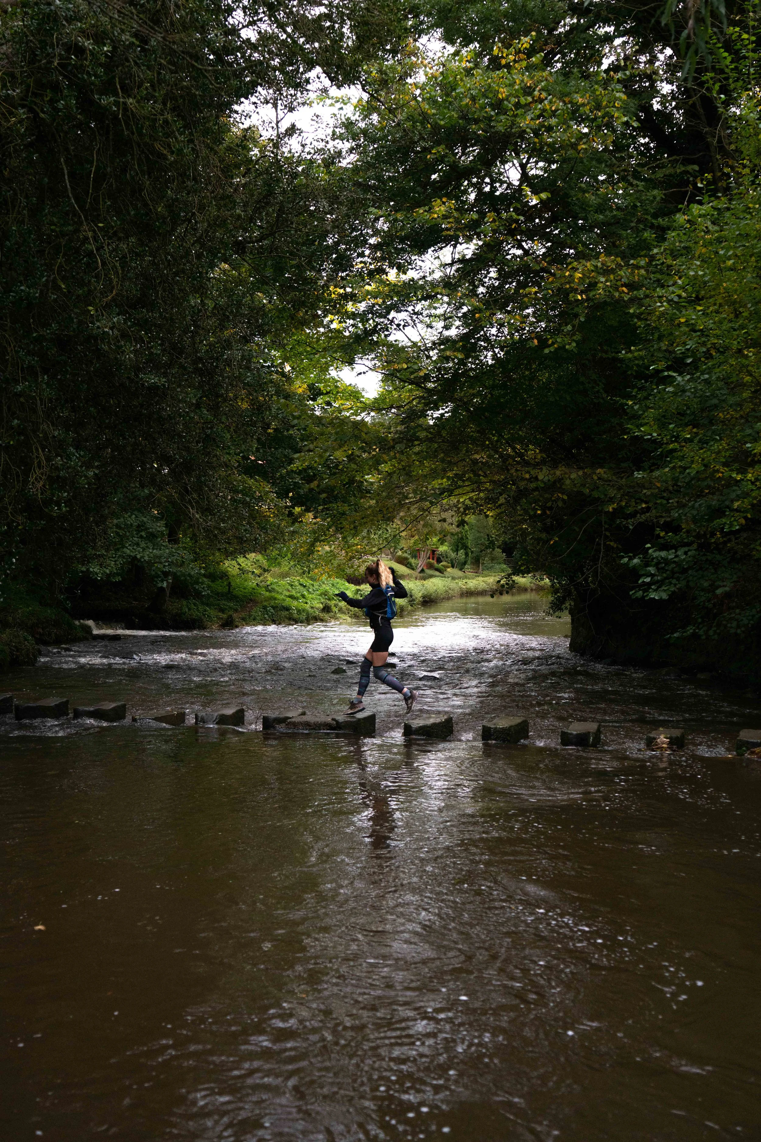 A person with a backpack crossing a small rocky step on a river in a wooded area with dense green trees and bushes.