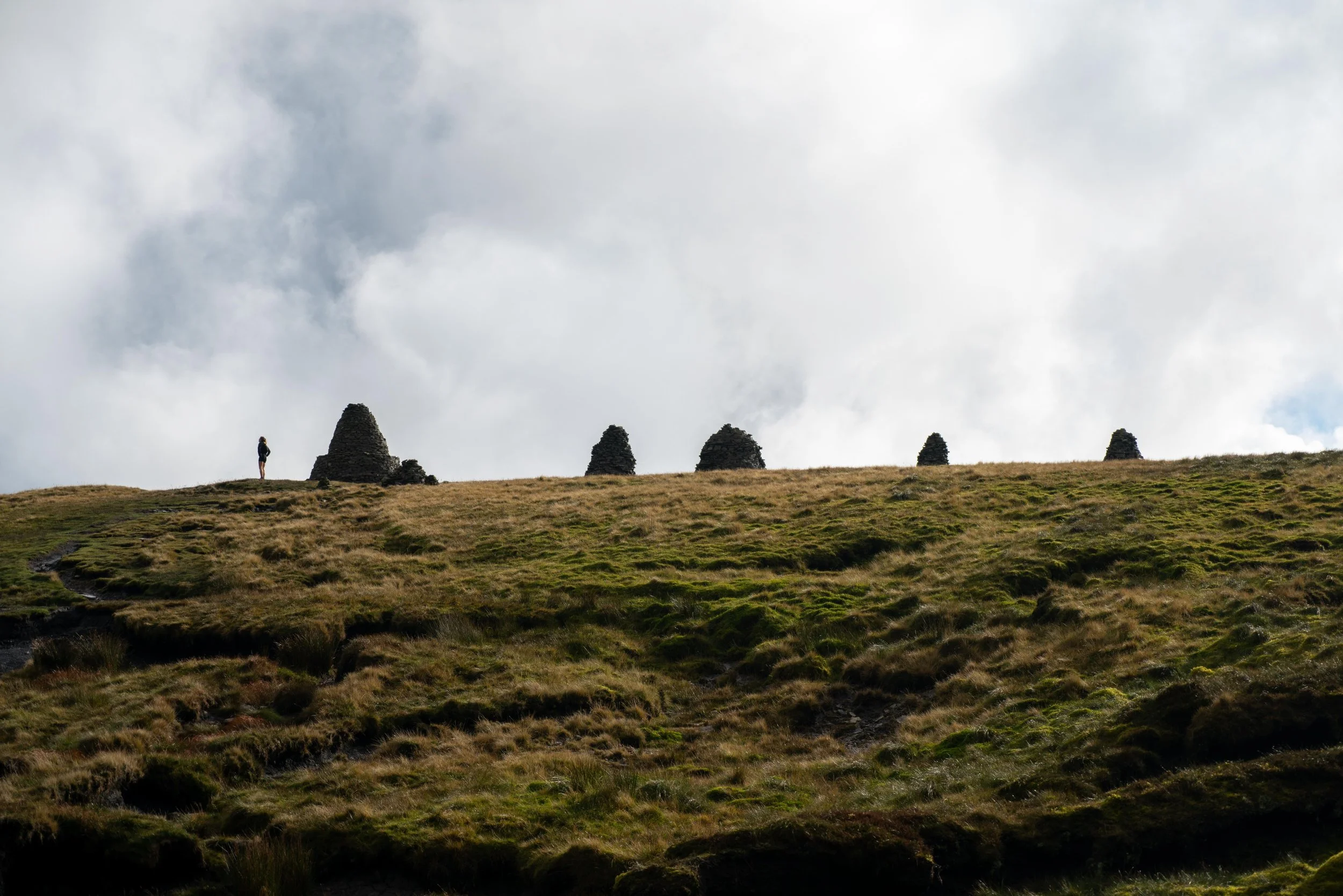 A person standing on a grassy hill with stone structures in the distance under a cloudy sky.