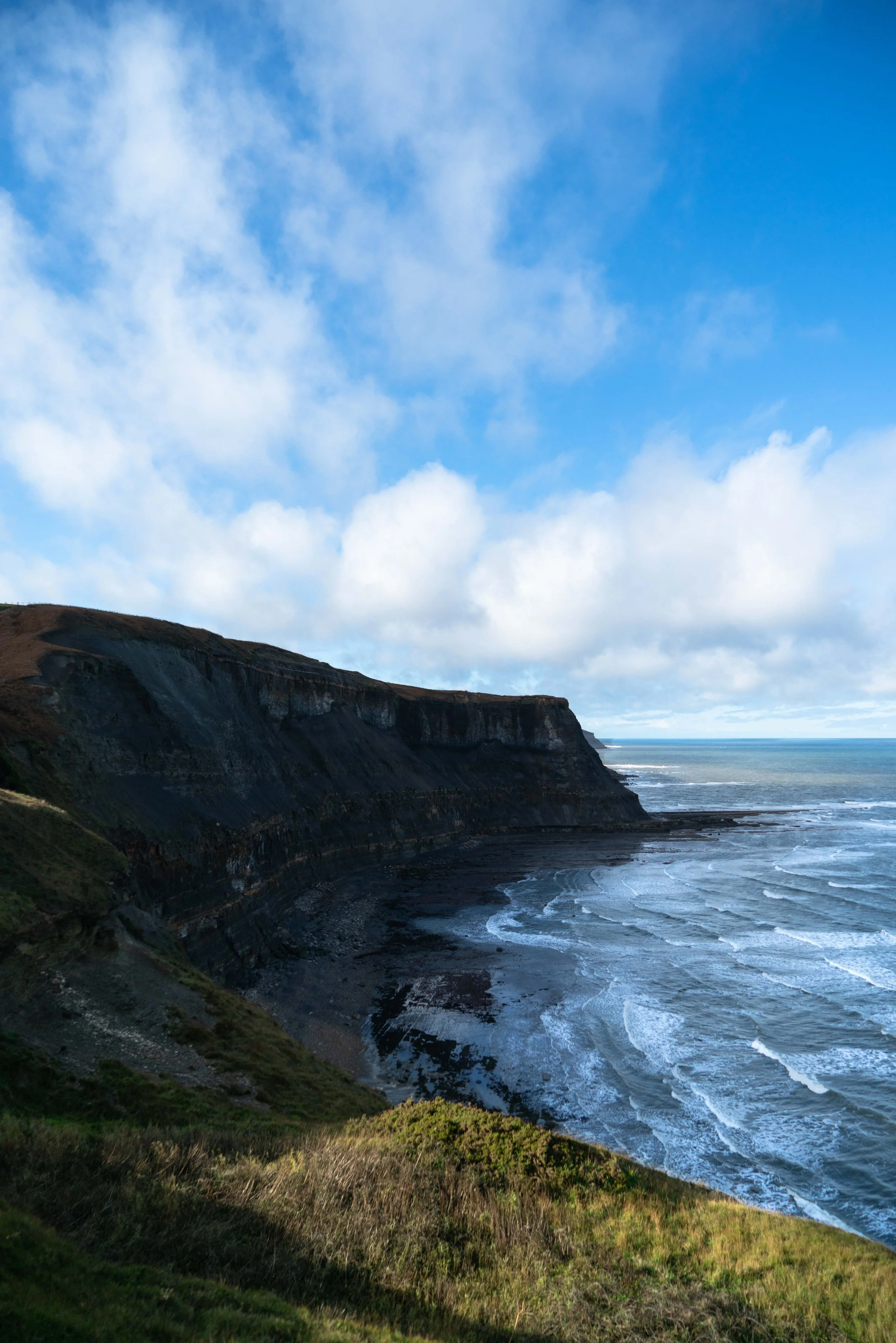 Cliffs along a coastline with the ocean below, under a blue sky with scattered clouds.