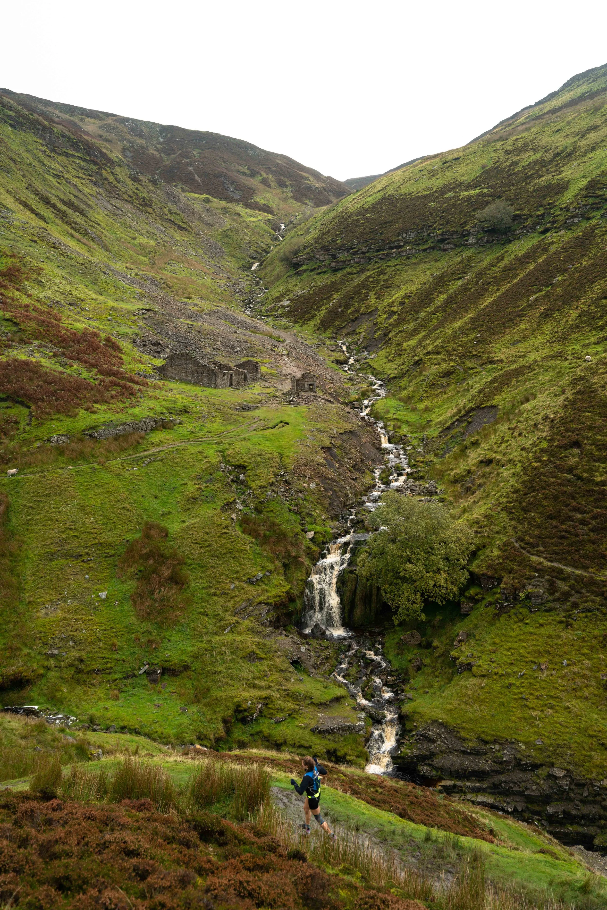 A person hiking along a grassy trail in a lush green valley with steep hills on either side. A small waterfall flows down the hillside into a stream below. The sky is overcast.