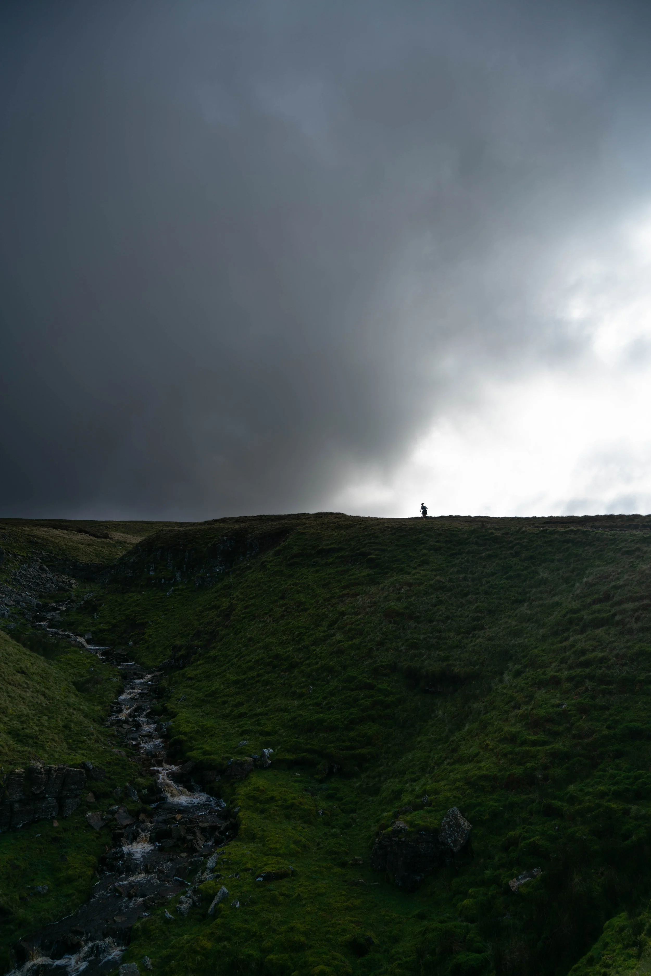 A small figure walking on a grassy hill under dark stormy clouds with a stream flowing down the hillside.
