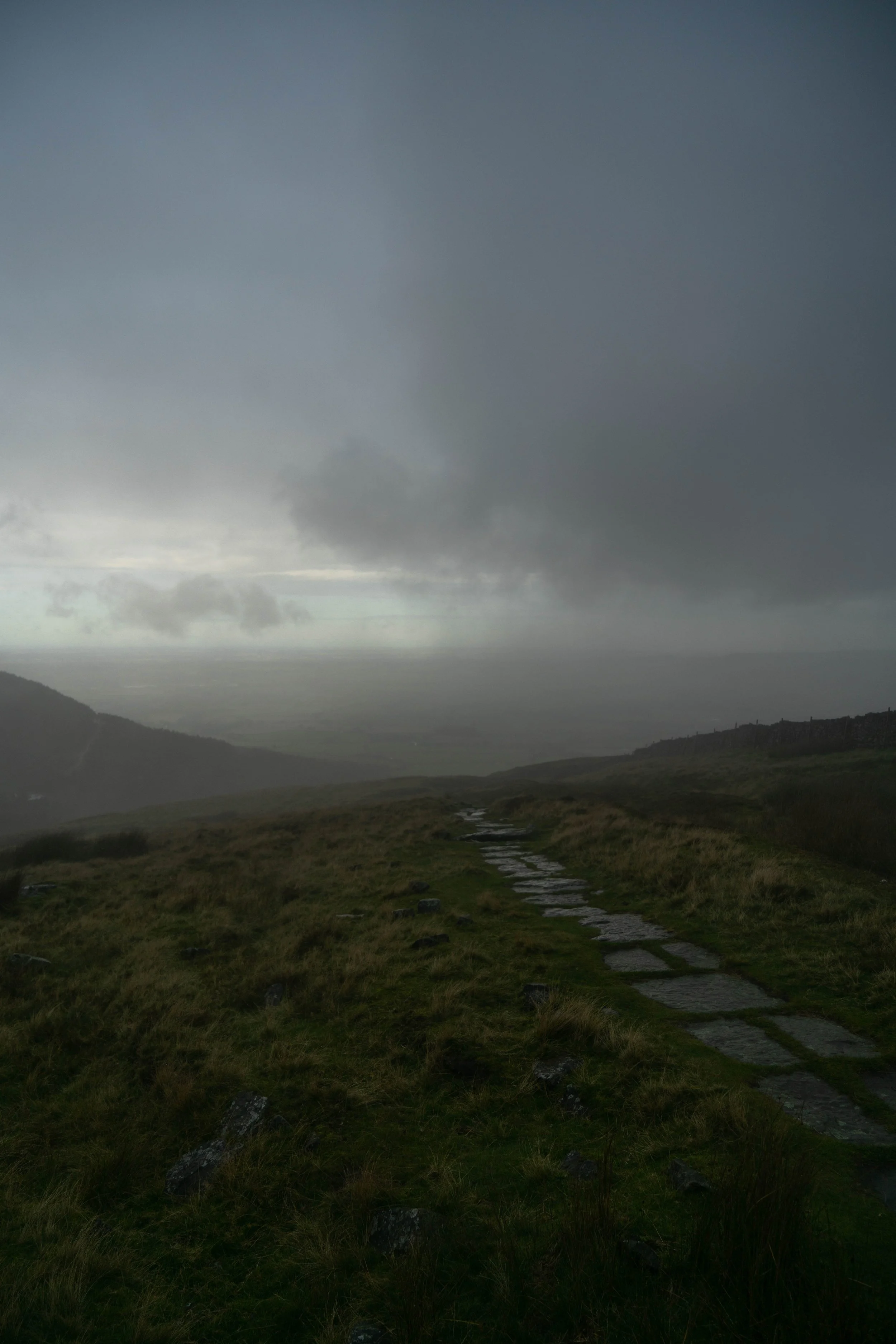 A landscape view of a grassy hillside leading to a distant horizon under a dark, cloudy sky, with a narrow stone path running through the grass.