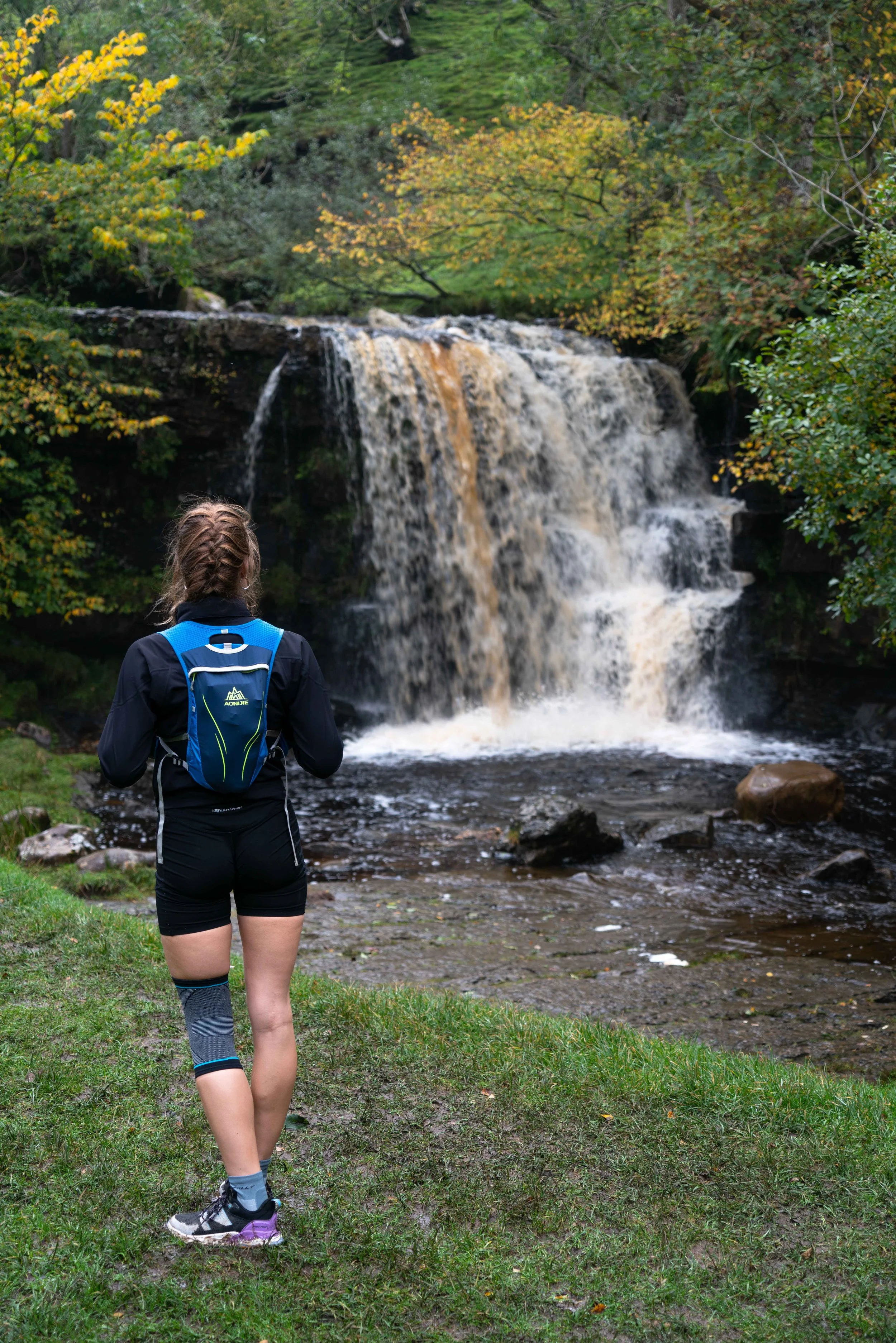 A woman in athletic gear with a blue backpack standing near a waterfall in a forest.