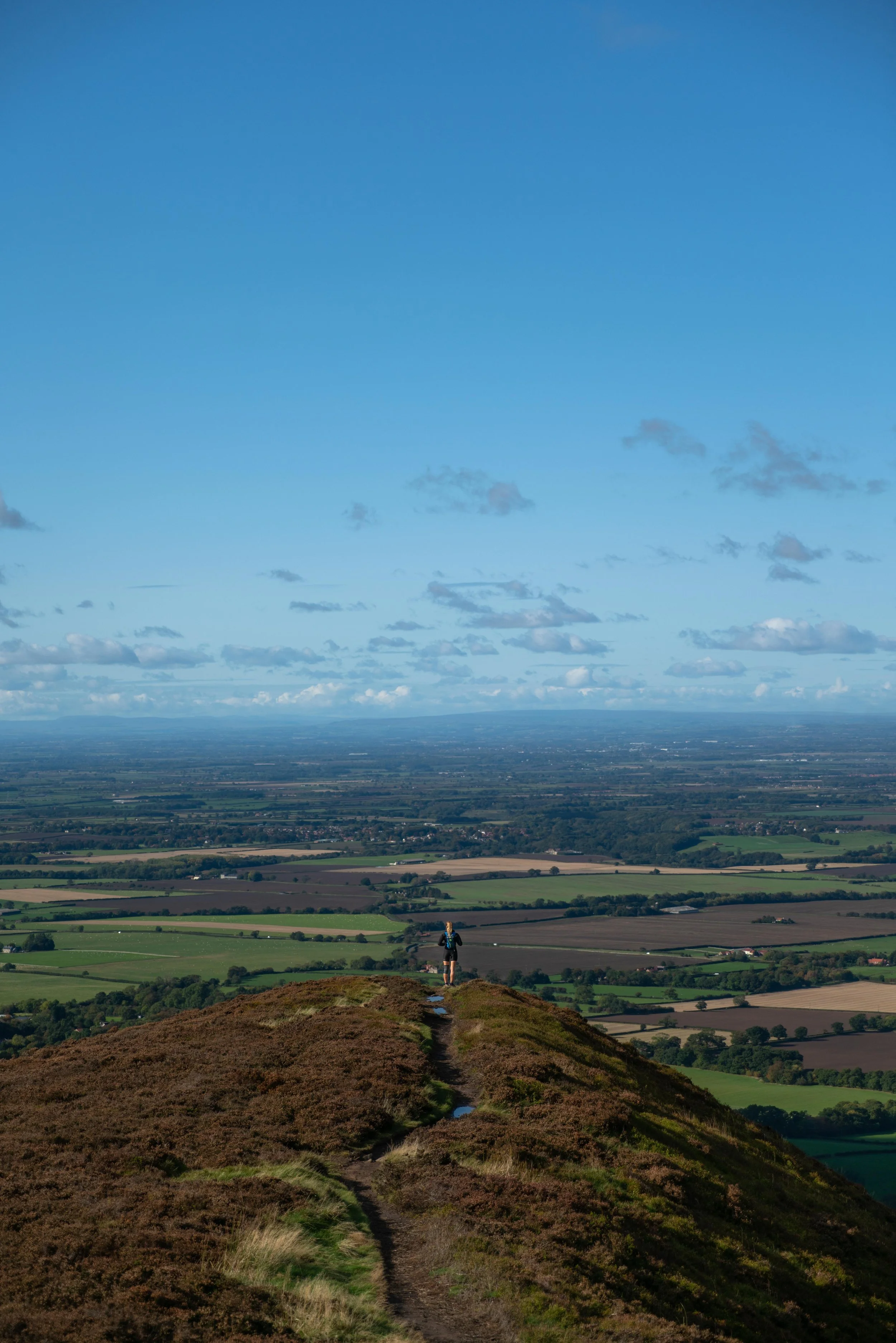 A person hiking on a trail atop a hill or mountain, overlooking a vast green and farmland landscape with scattered clouds and a blue sky.