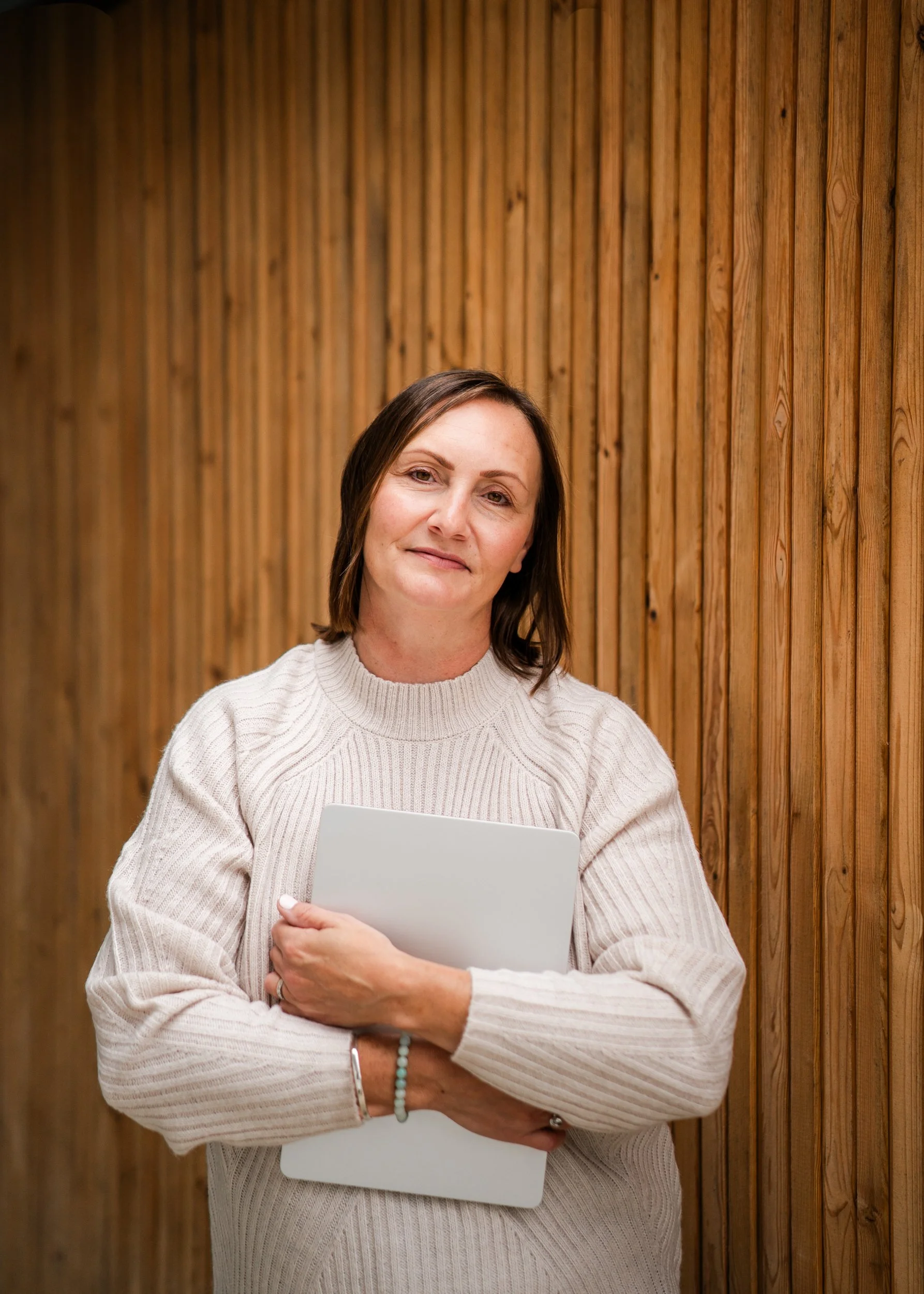 A woman with shoulder-length brown hair and a light sweater holding a closed laptop and standing in front of a wooden wall.