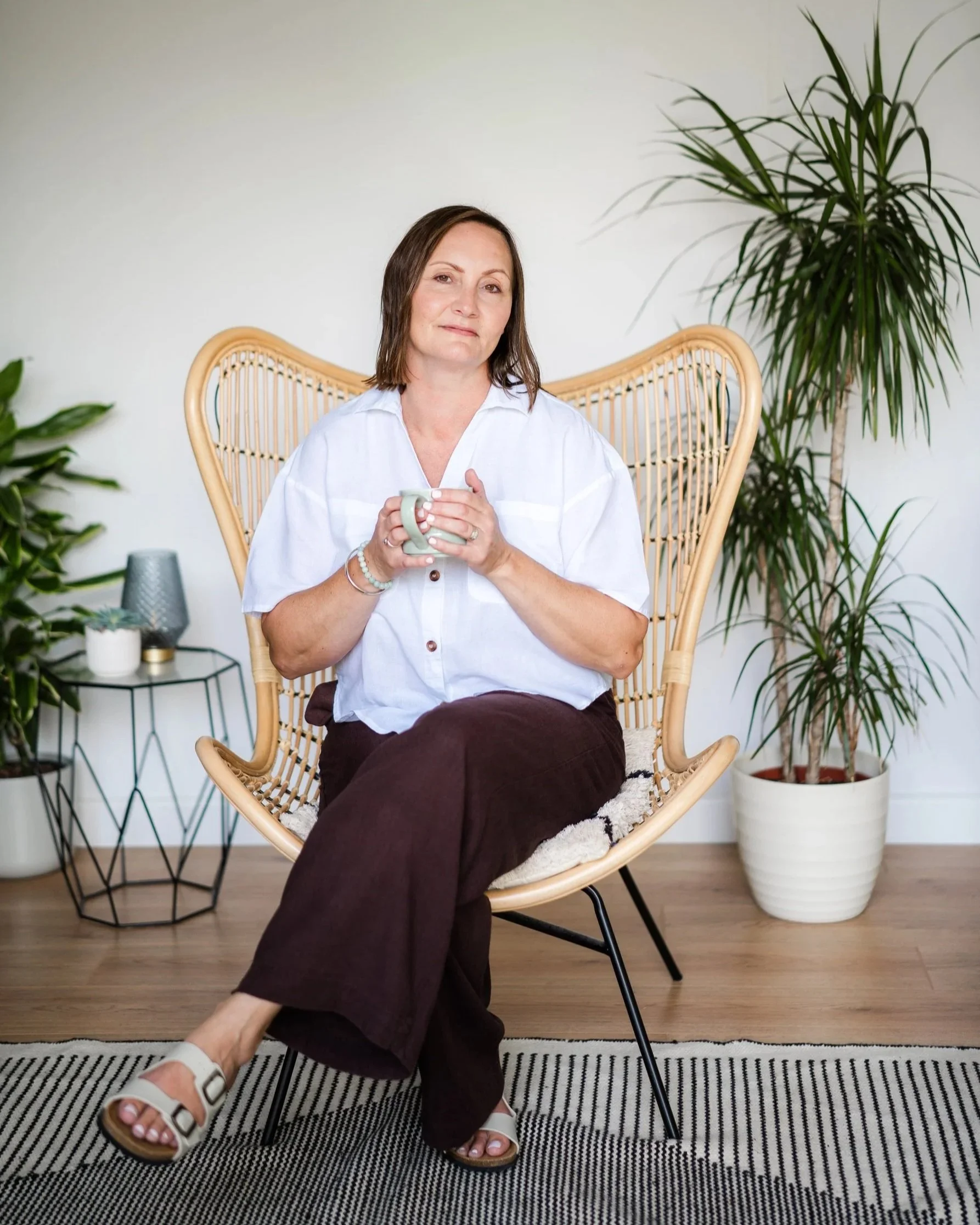 A woman sitting in a wicker chair holding a mug, with plants and side tables in a minimalistic and modern living space.