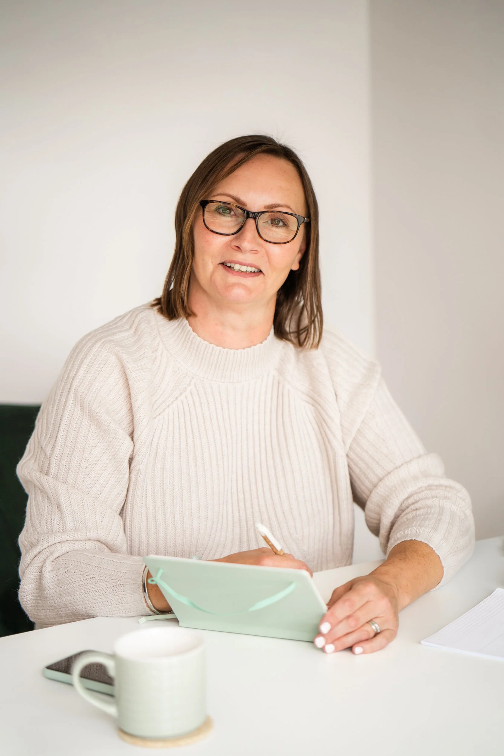 A woman with glasses sitting at a white desk, writing in a notebook, with a mint green box, a mug, a smartphone, and a notepad on the desk.