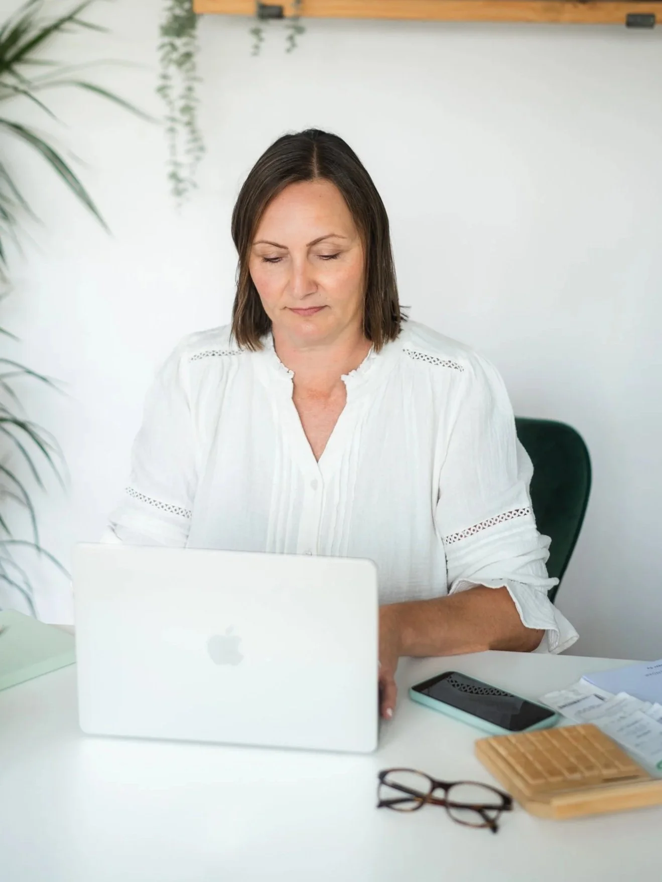A woman sitting at a white desk with a silver MacBook, a smartphone, reading glasses, and papers, working in an office environment with a white wall and a plant partially visible.