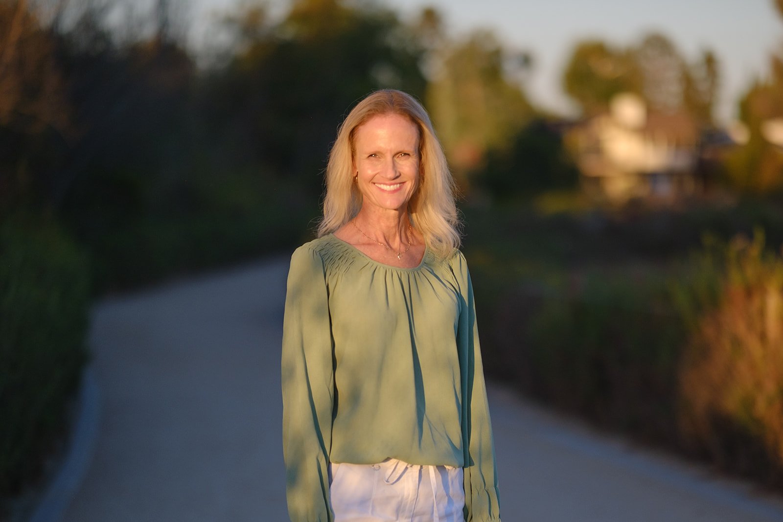 A woman smiling outdoors on a path during sunset, wearing a light green blouse and white pants.
