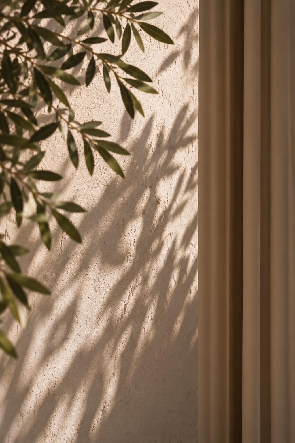 Shadows of green leaves cast on a textured beige wall next to beige vertical slats.