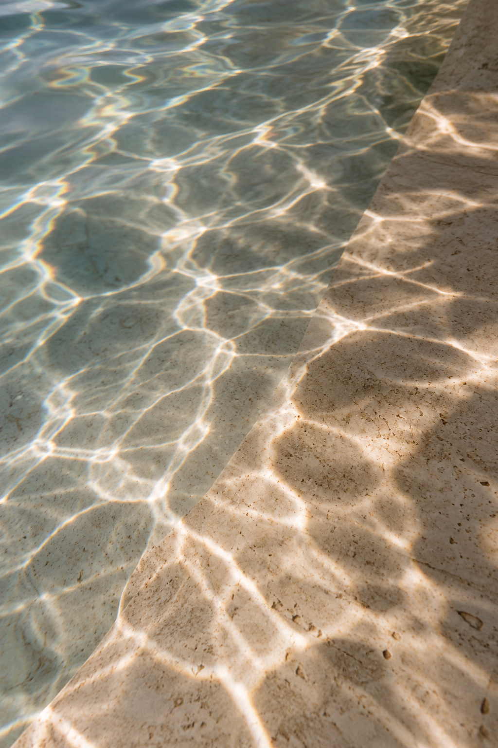 Sunlit water with shimmering reflections casting patterns onto a sandy surface along a shoreline.