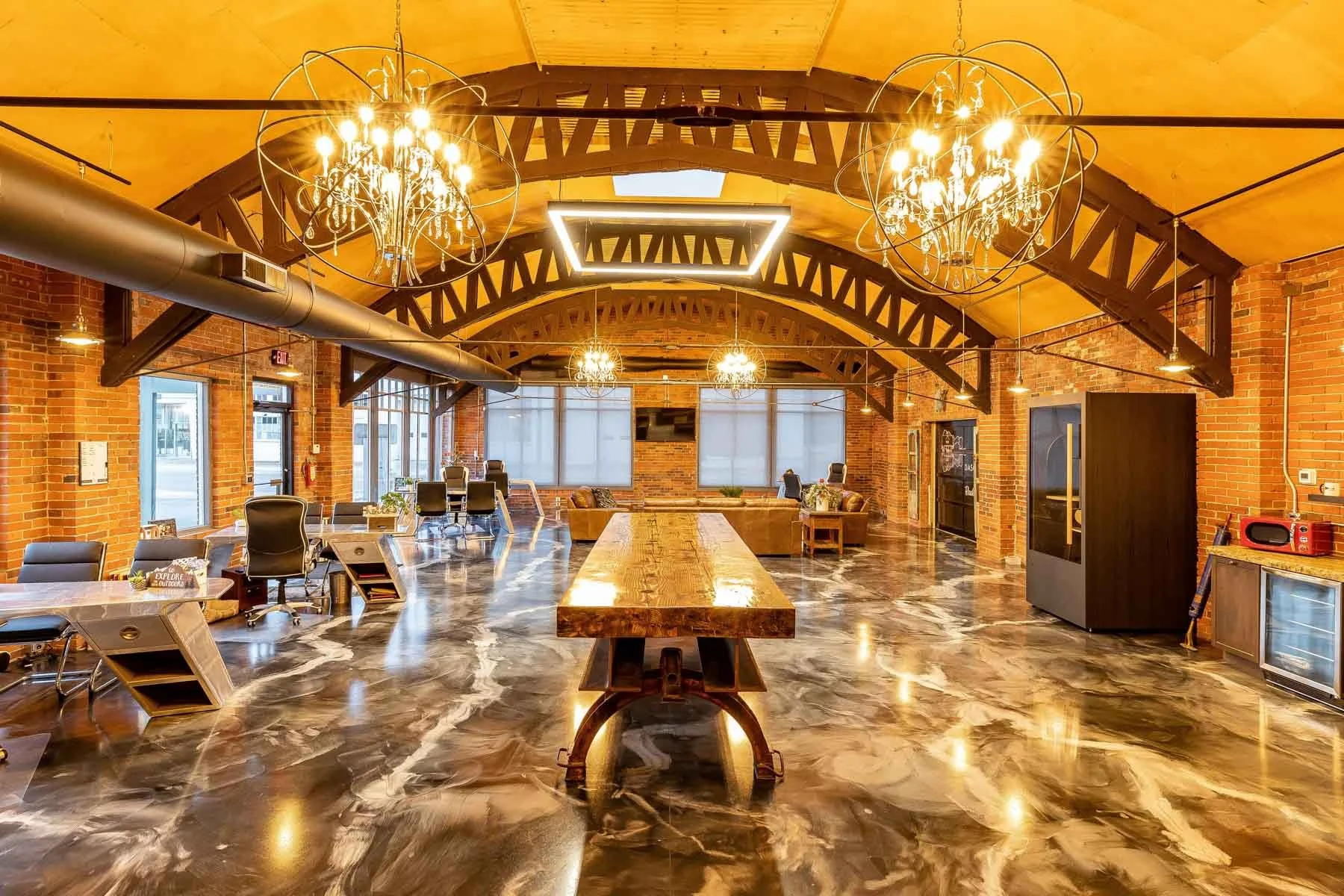 Interior of a modern, spacious office with exposed brick walls, high wooden ceilings, and polished marble floors. The room features a large wooden table in the center, multiple chairs, and decorative chandeliers hanging from the ceiling. There are large windows at the back allowing natural light, along with various seating areas and office equipment.
