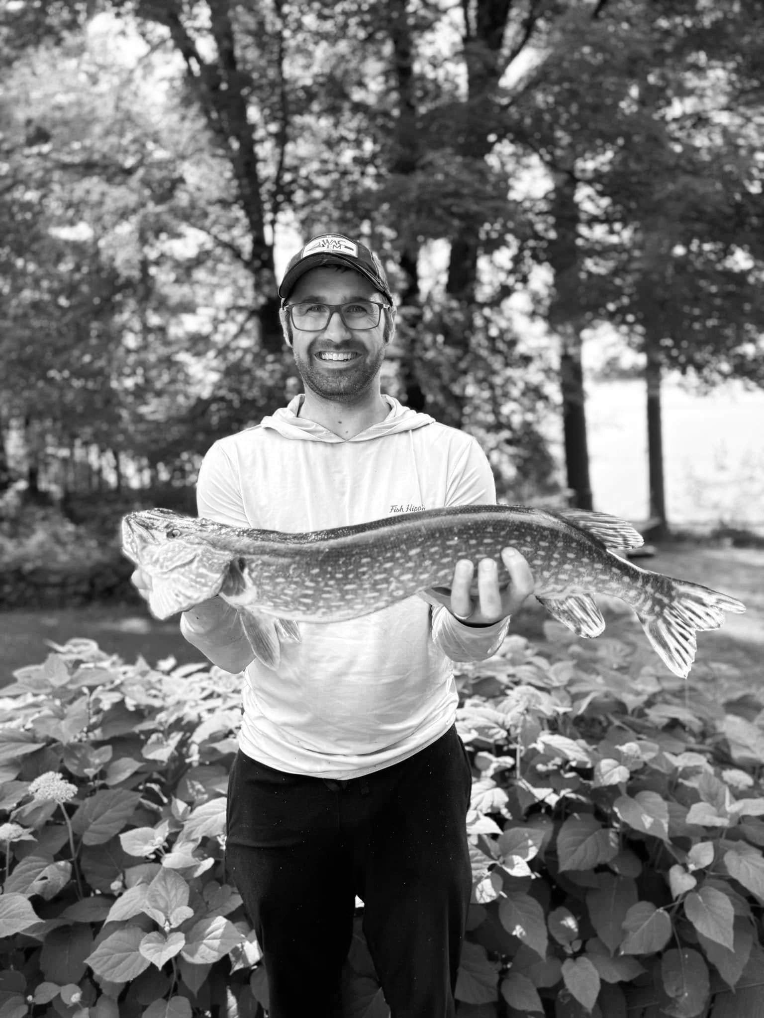 A man standing outdoors in front of trees, holding a large fish with both hands, smiling at the camera.