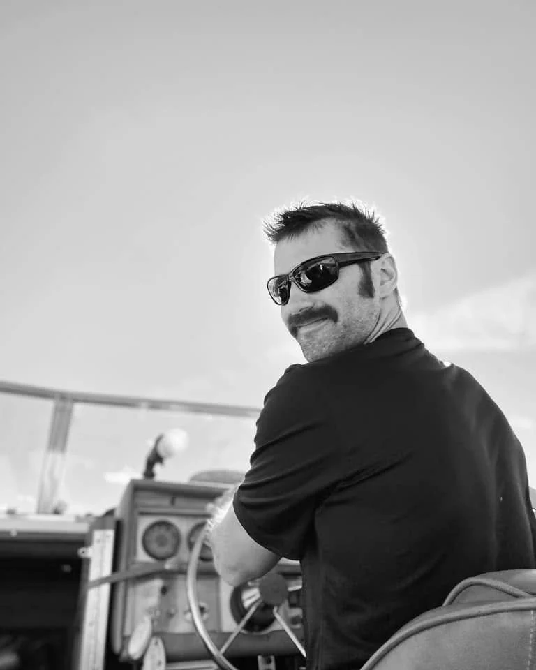 A man with a mustache and sunglasses sitting at a steering wheel on a boat, looking back and smiling.